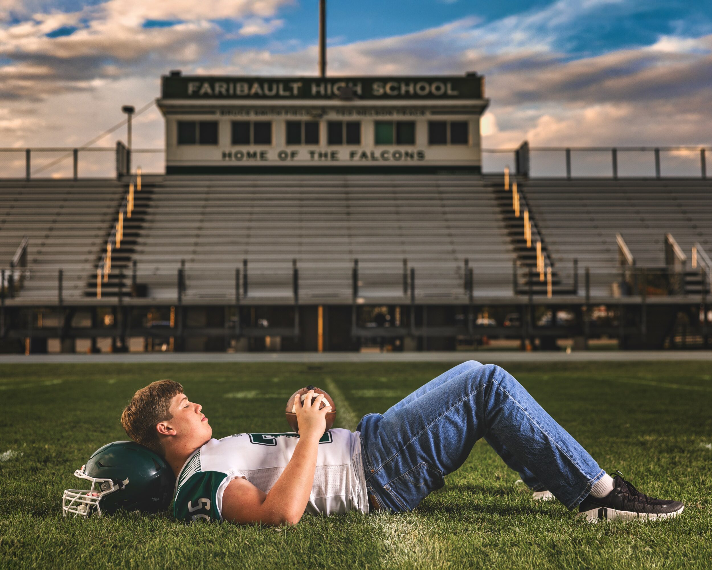 A football player lies on the field holding a football, with empty bleachers and a school sign in the background.