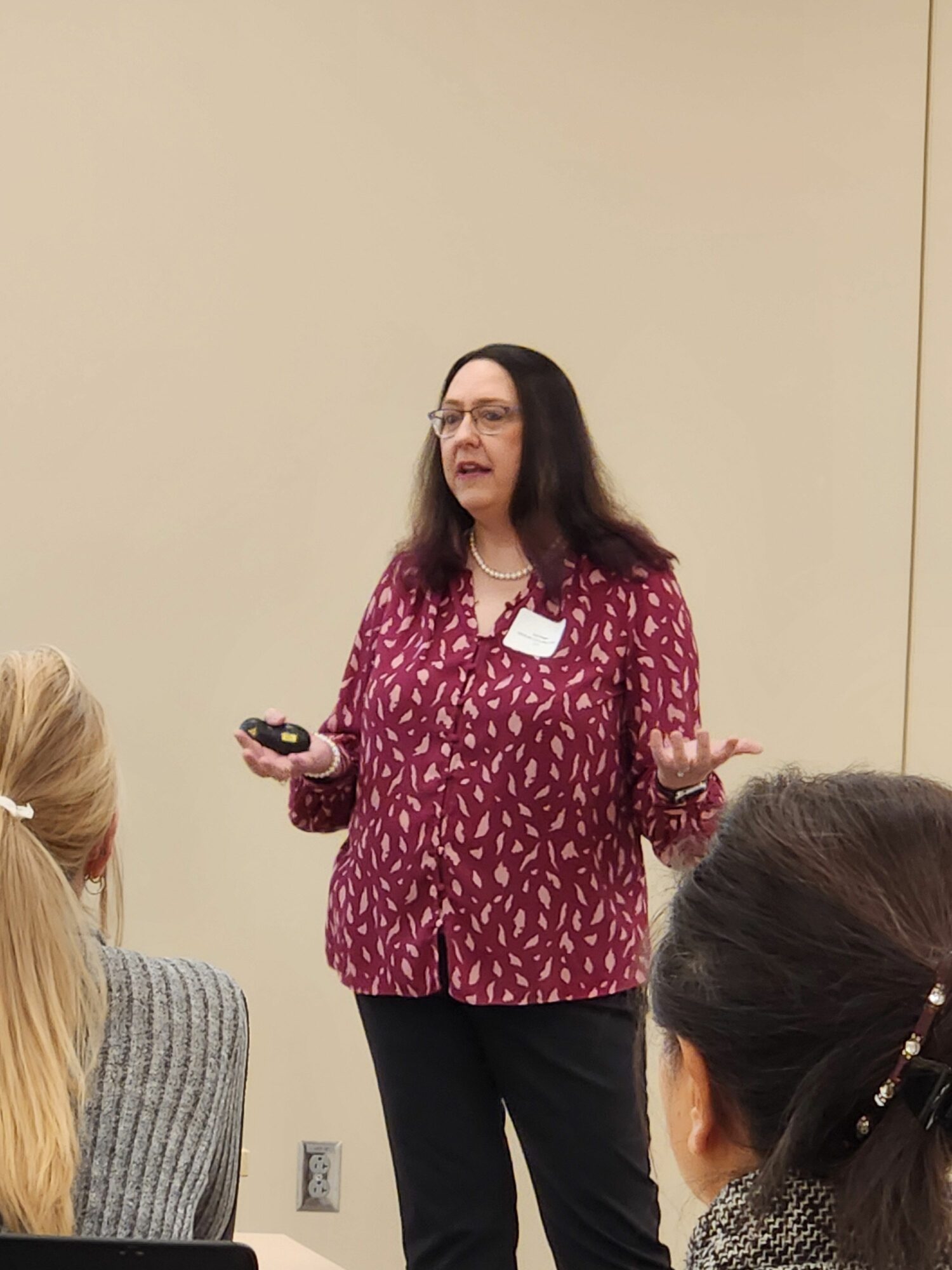 Woman with long dark hair wearing a red patterned blouse and black pants, holding a phone, standing indoors.