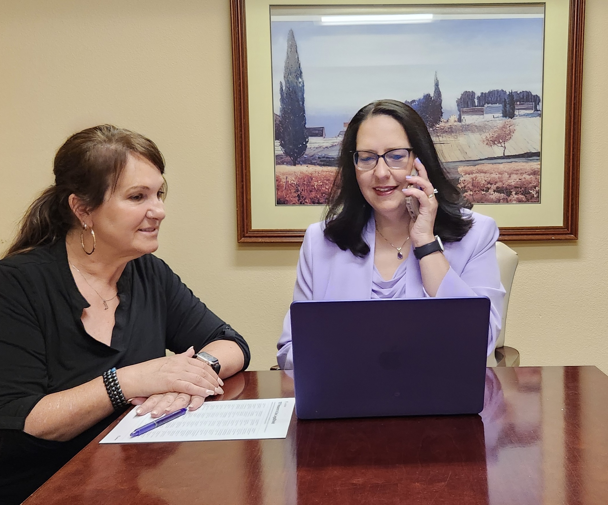 Two women sit at a table with a laptop, one smiling and the other talking, in an office setting.