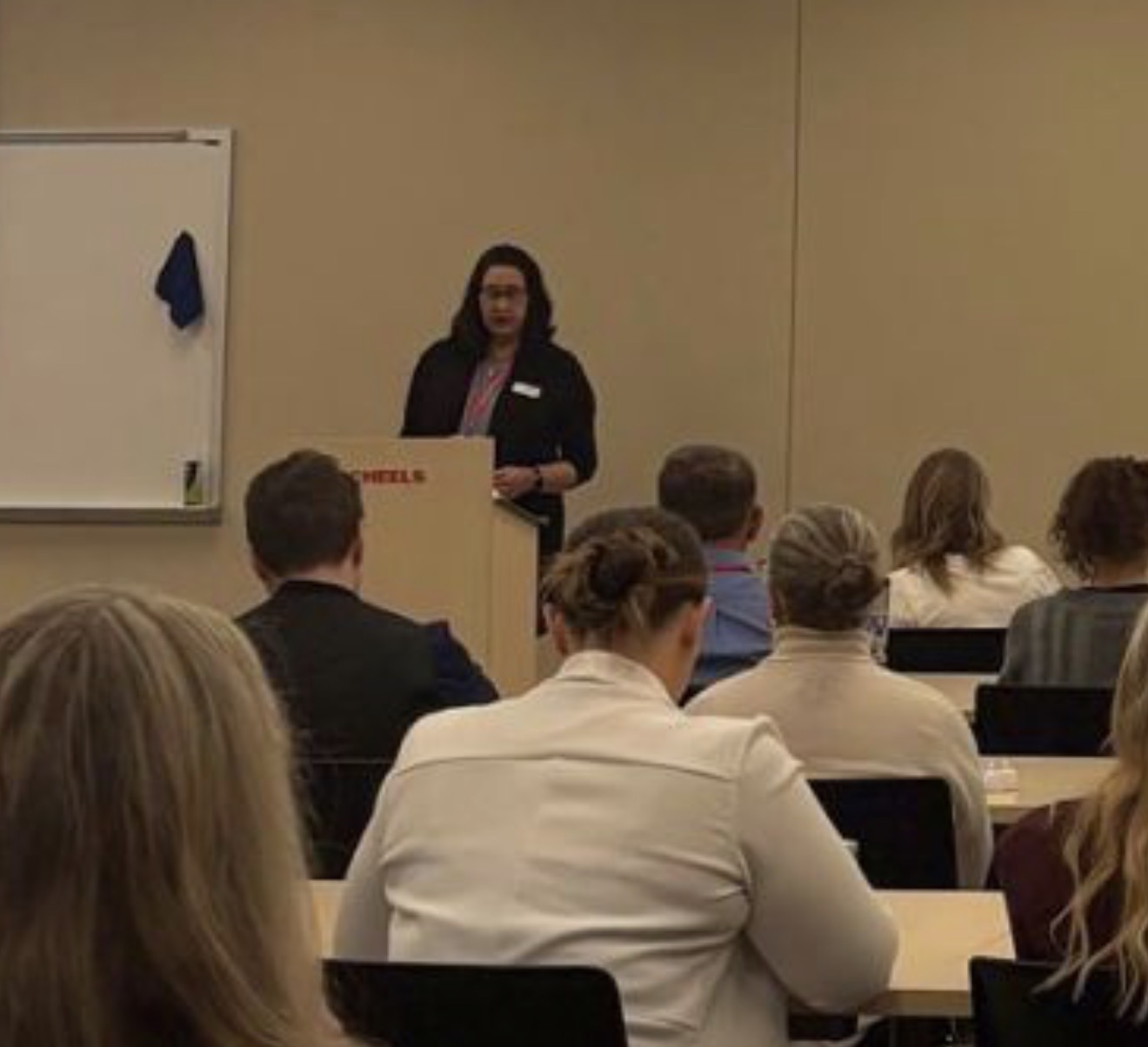 Woman standing at podium giving a presentation to seated audience in a classroom setting.