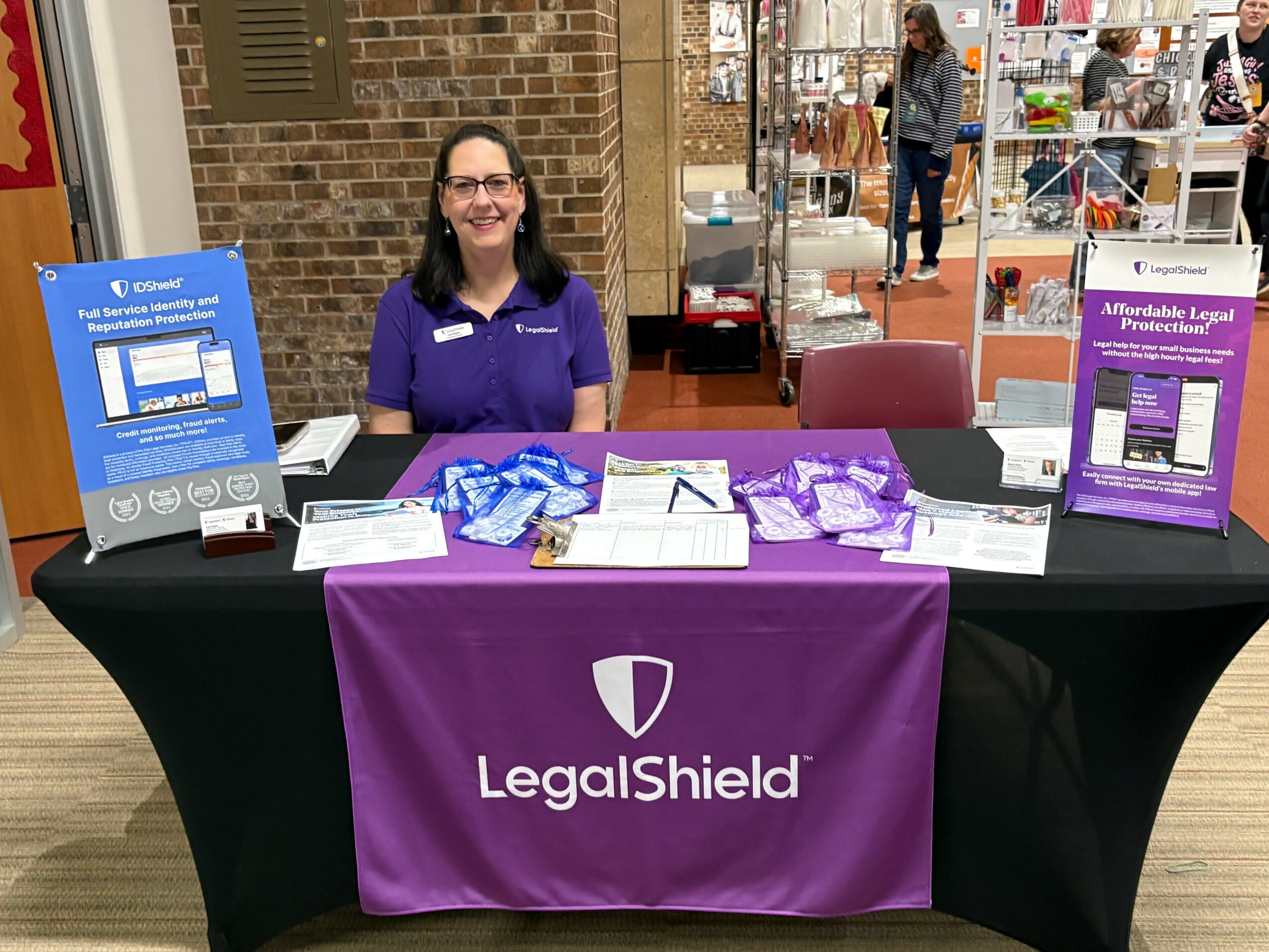 Woman sitting at a table with LegalShield promotional materials, in a public indoor space, smiling, wearing glasses and a purple shirt.