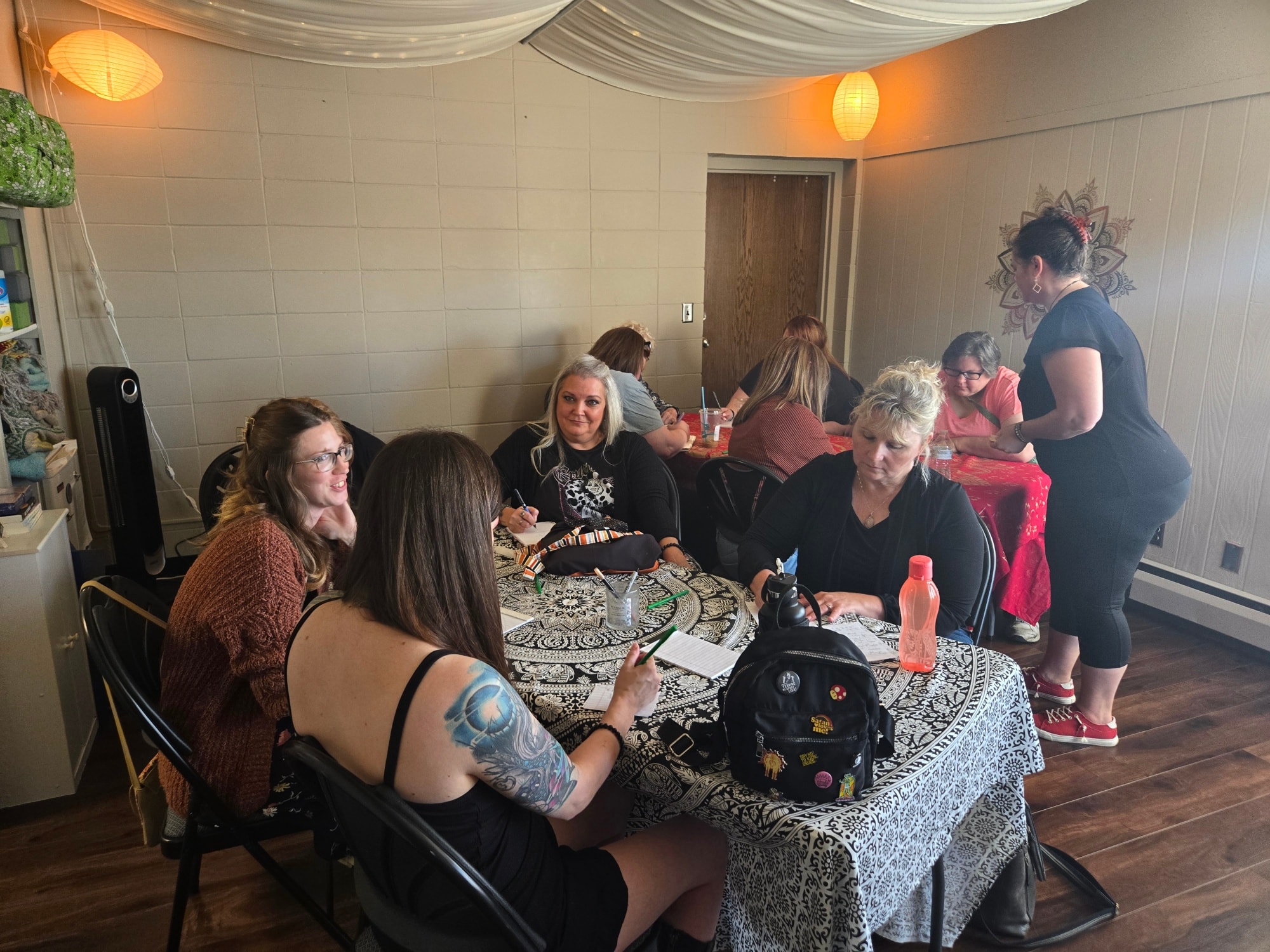 Group of women sitting around a table in a room with wooden floor and beige walls, some women are talking and using phones.