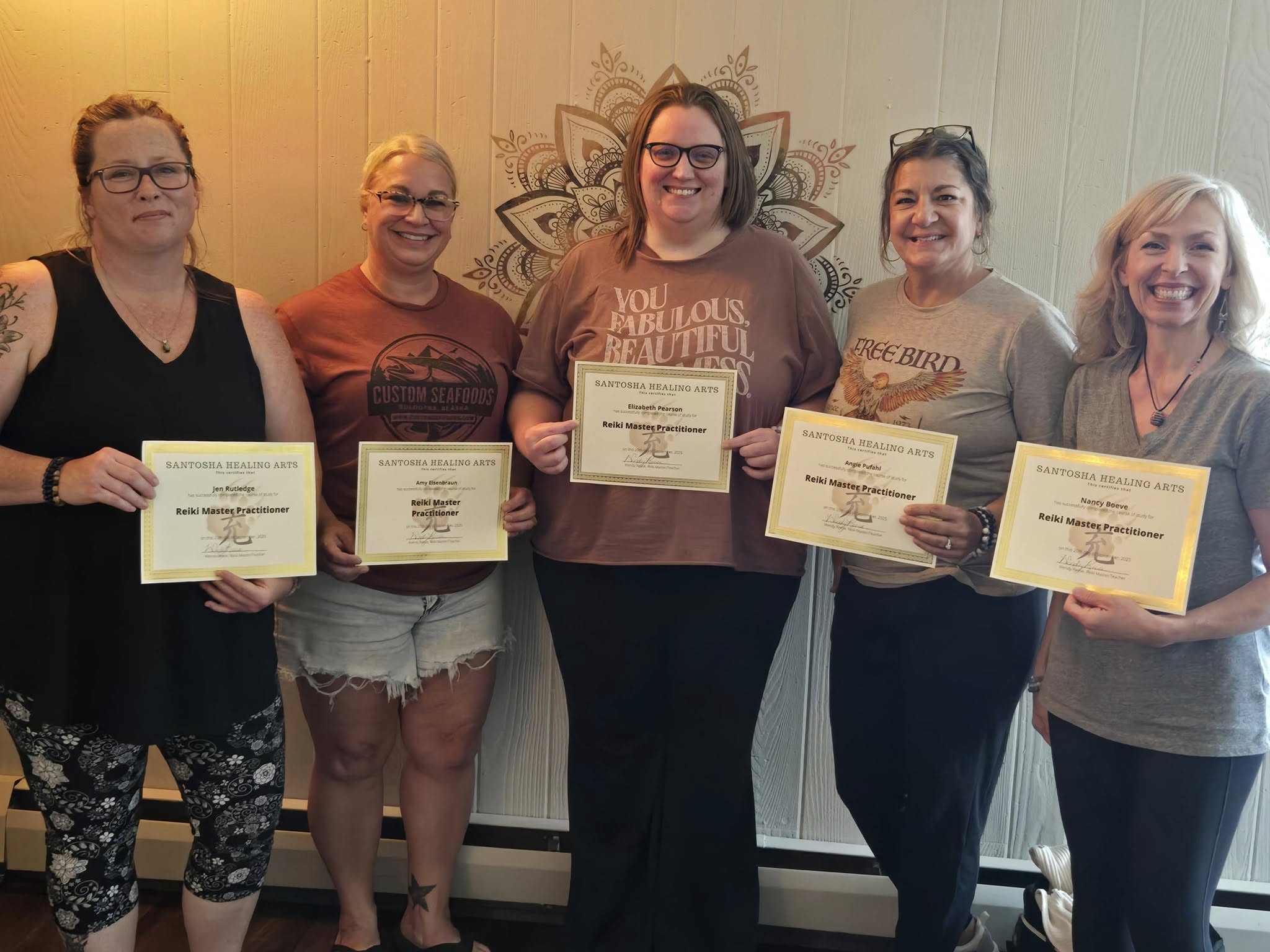 Five women standing indoors, holding certificates, smiling, with a decorative wall behind them.