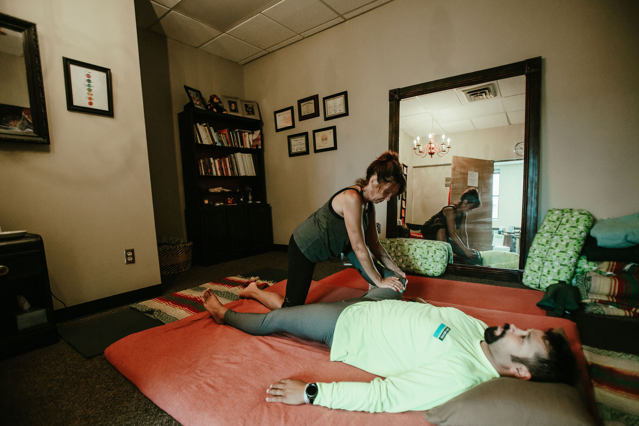 Person lying on their stomach on a bed, another person kneeling beside them, in a cozy room with a large mirror.