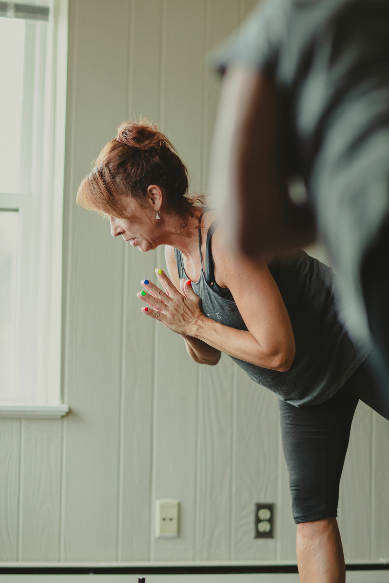 Woman practicing yoga indoors, leaning forward with hands pressed together near her face, near a window with natural light.