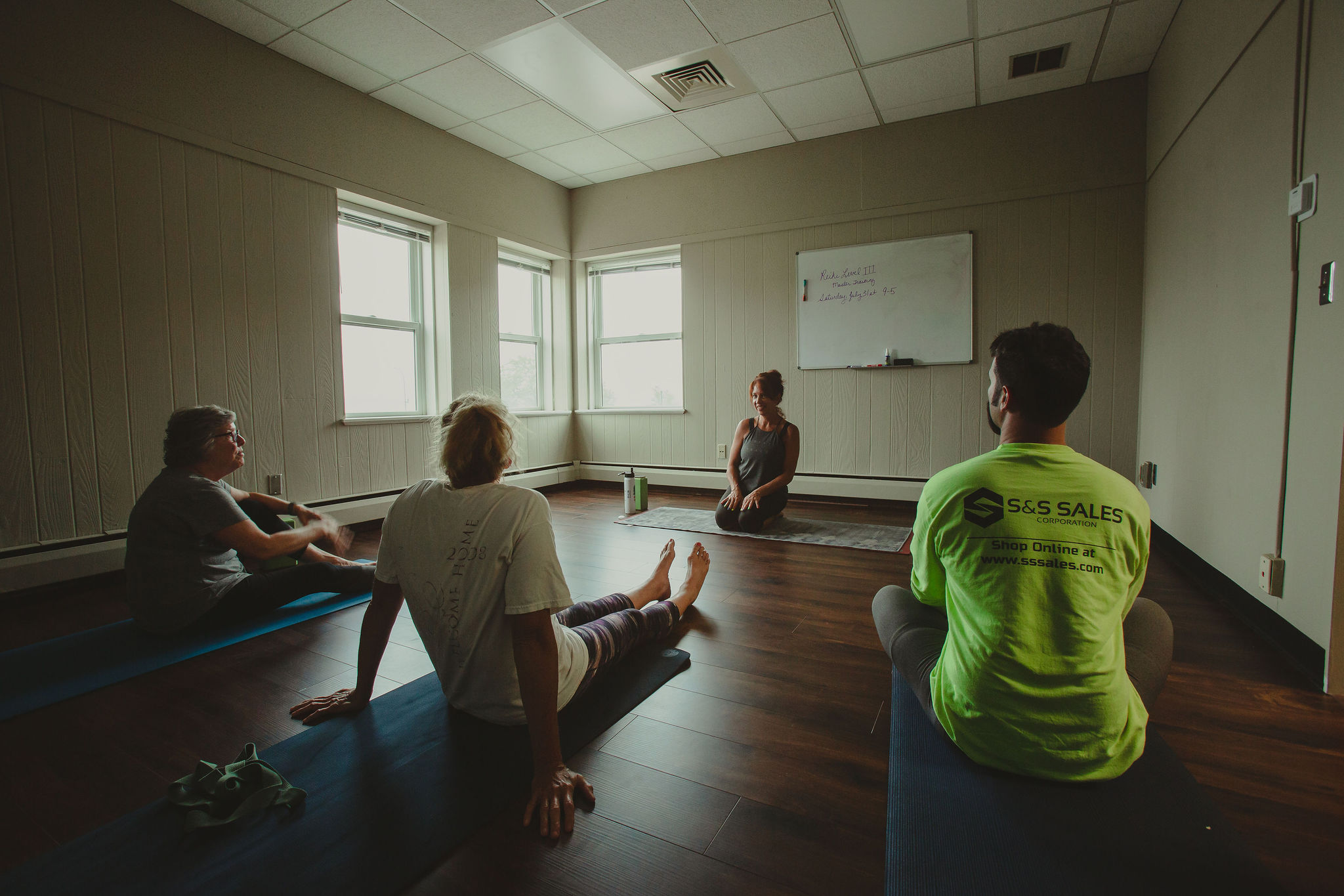 Four people sit on mats in a room with a whiteboard, listening to a woman at the front. Three windows let in natural light.