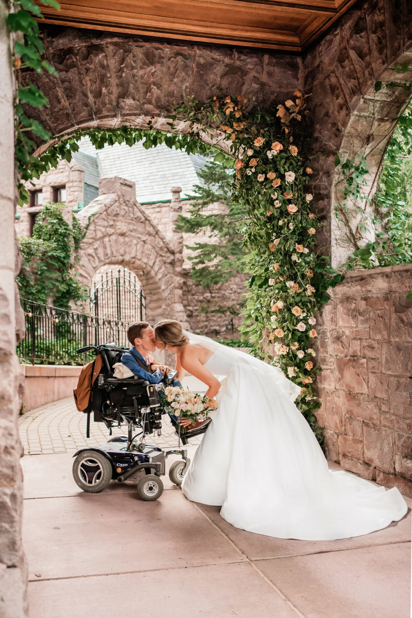 Bride in wedding dress leaning over wheelchair, kissing, under stone archway with flowers and greenery, outdoor setting.