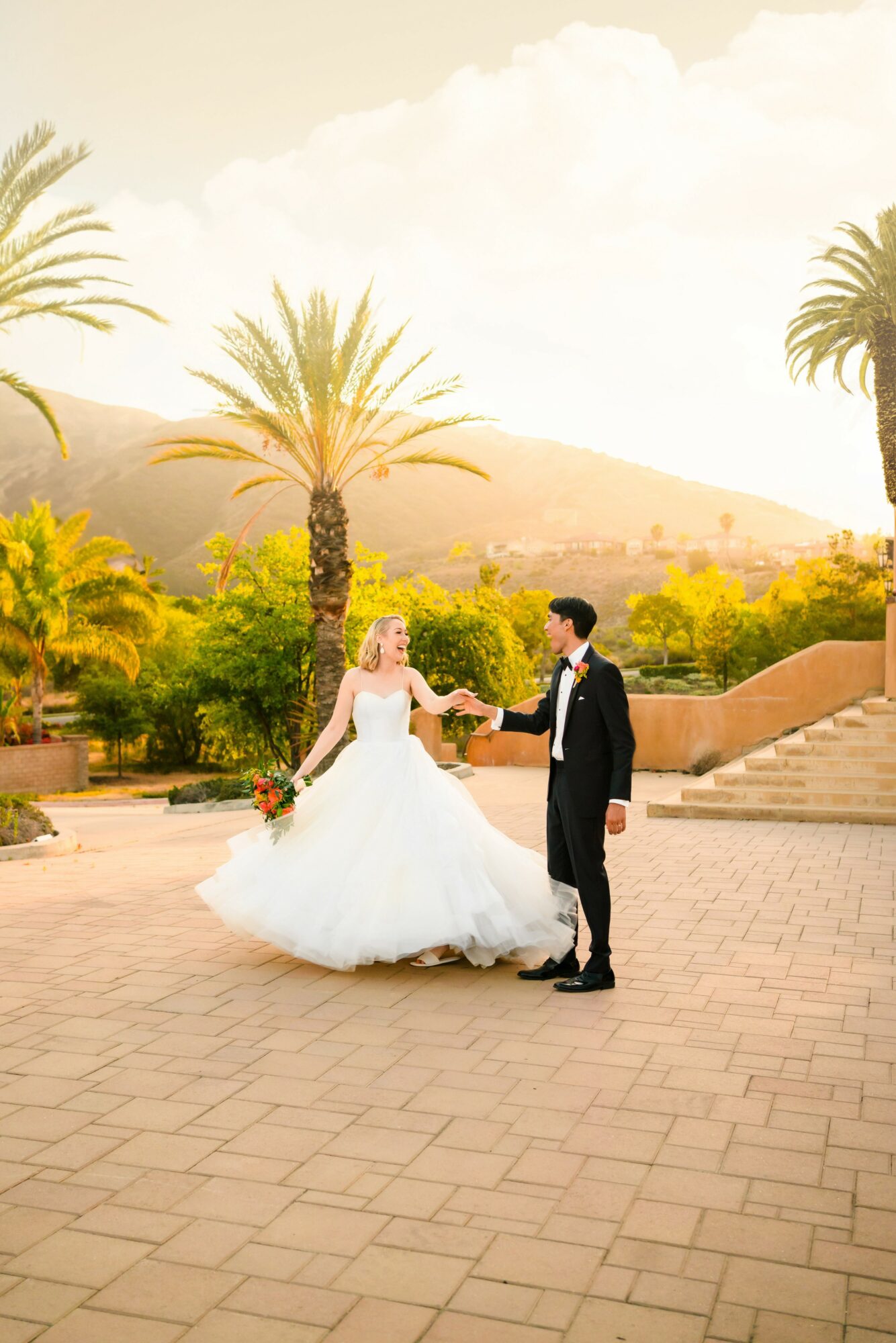 Bride and groom holding hands outdoors with palm trees and mountains in background, sunny sky, paved ground, wedding attire.