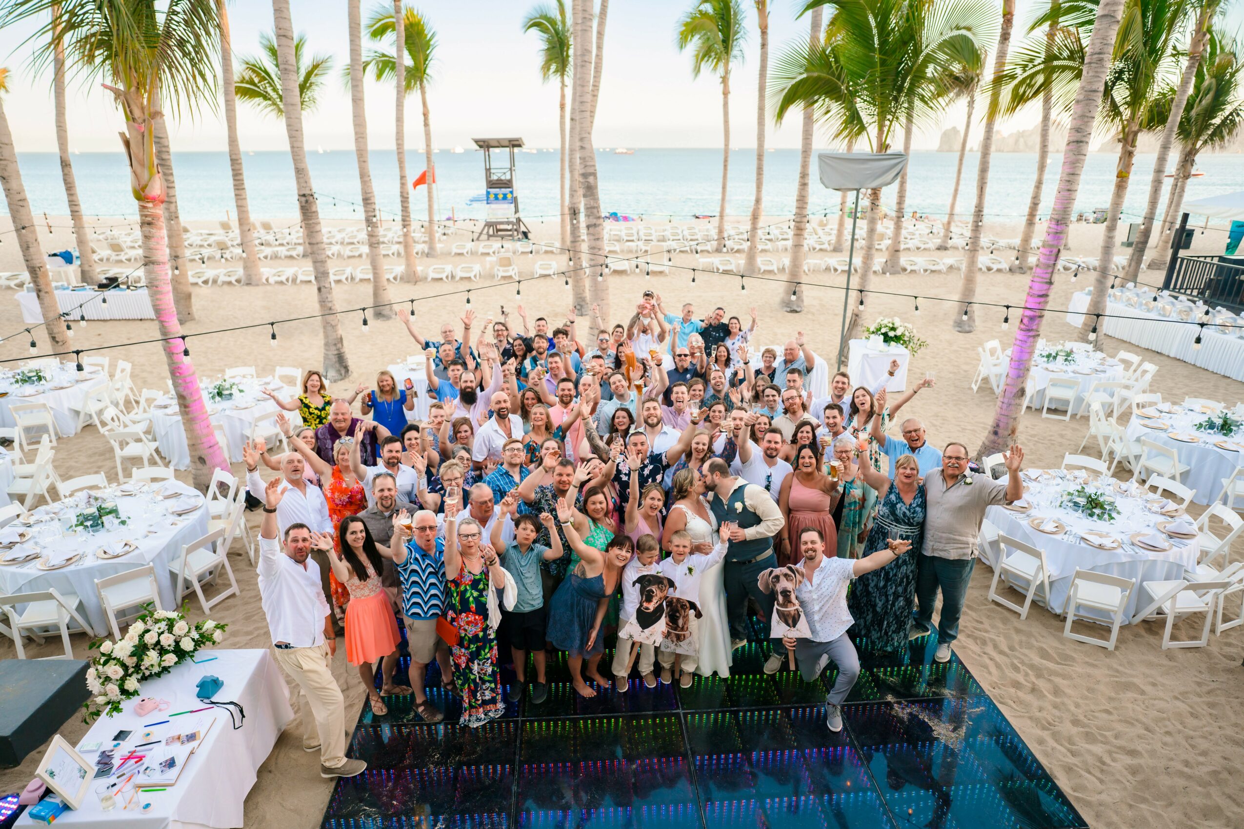 Large group of people gathered on a beach with palm trees, tables, and ocean in the background, celebrating outdoors.