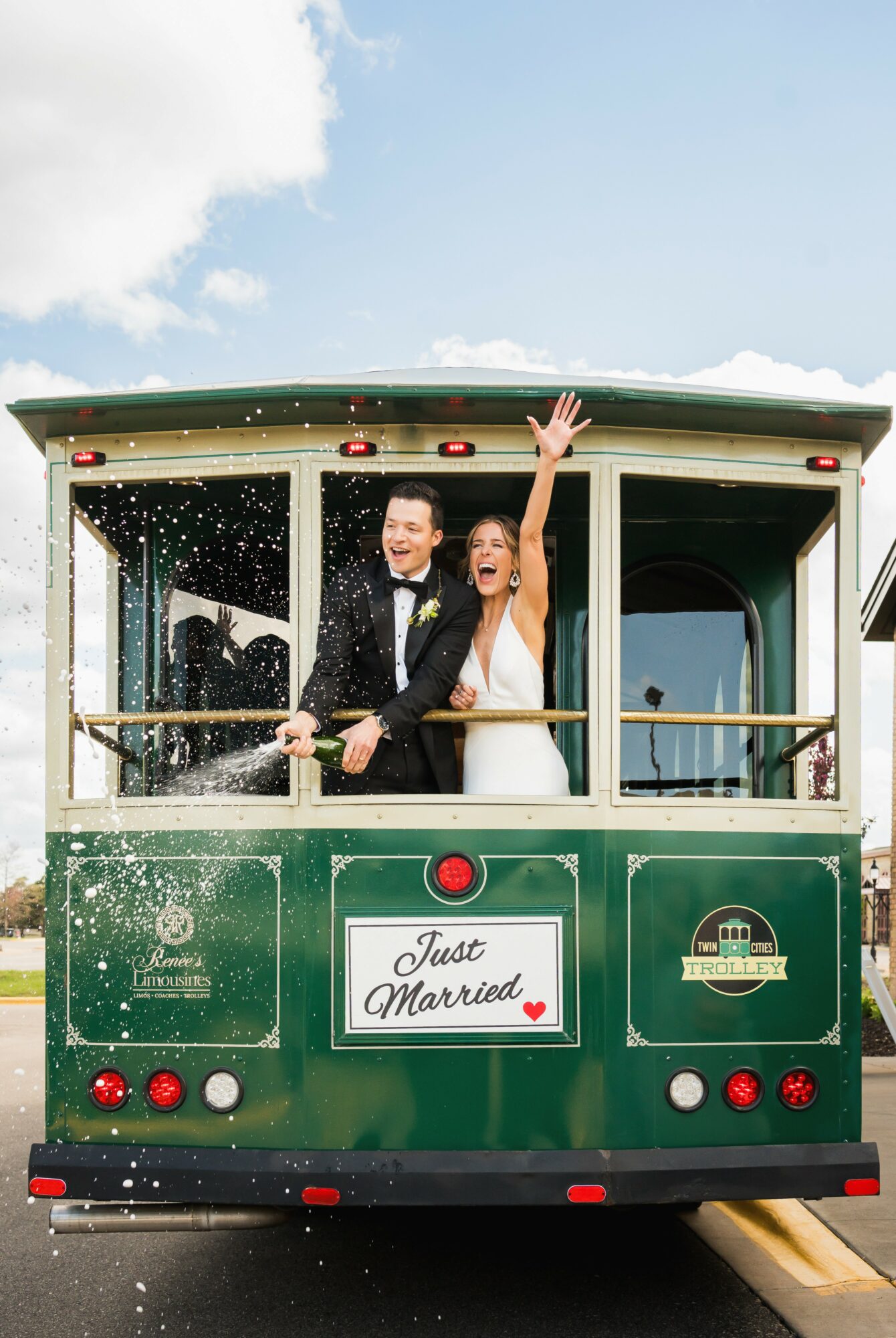A newlywed couple celebrating on a green trolley with a 'Just Married' sign, waving and smiling, with water splashing around.
