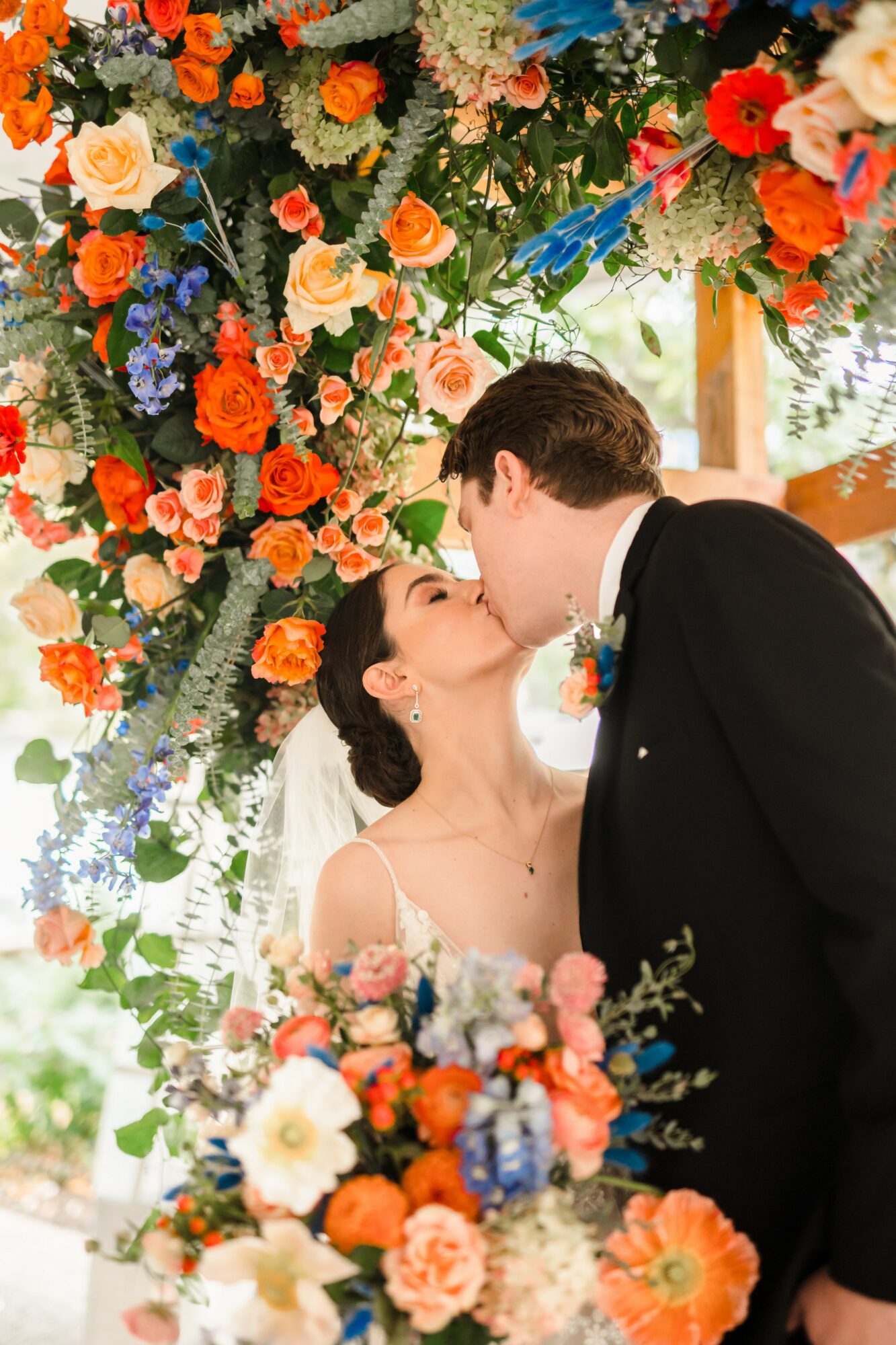 A couple kissing under a colorful floral arch at a wedding, with flowers in the foreground and background.