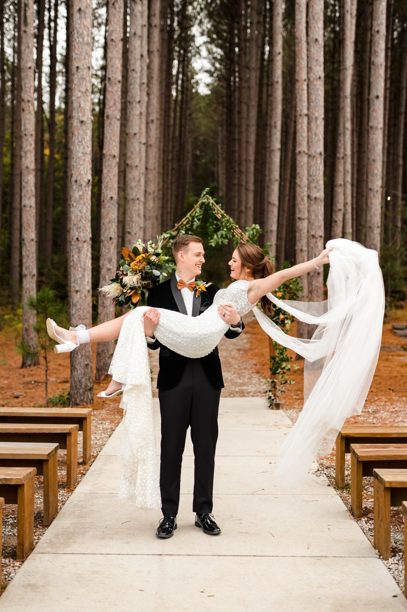 A couple in wedding attire standing on a forest path, with the groom holding the bride, surrounded by tall trees and wooden benches.