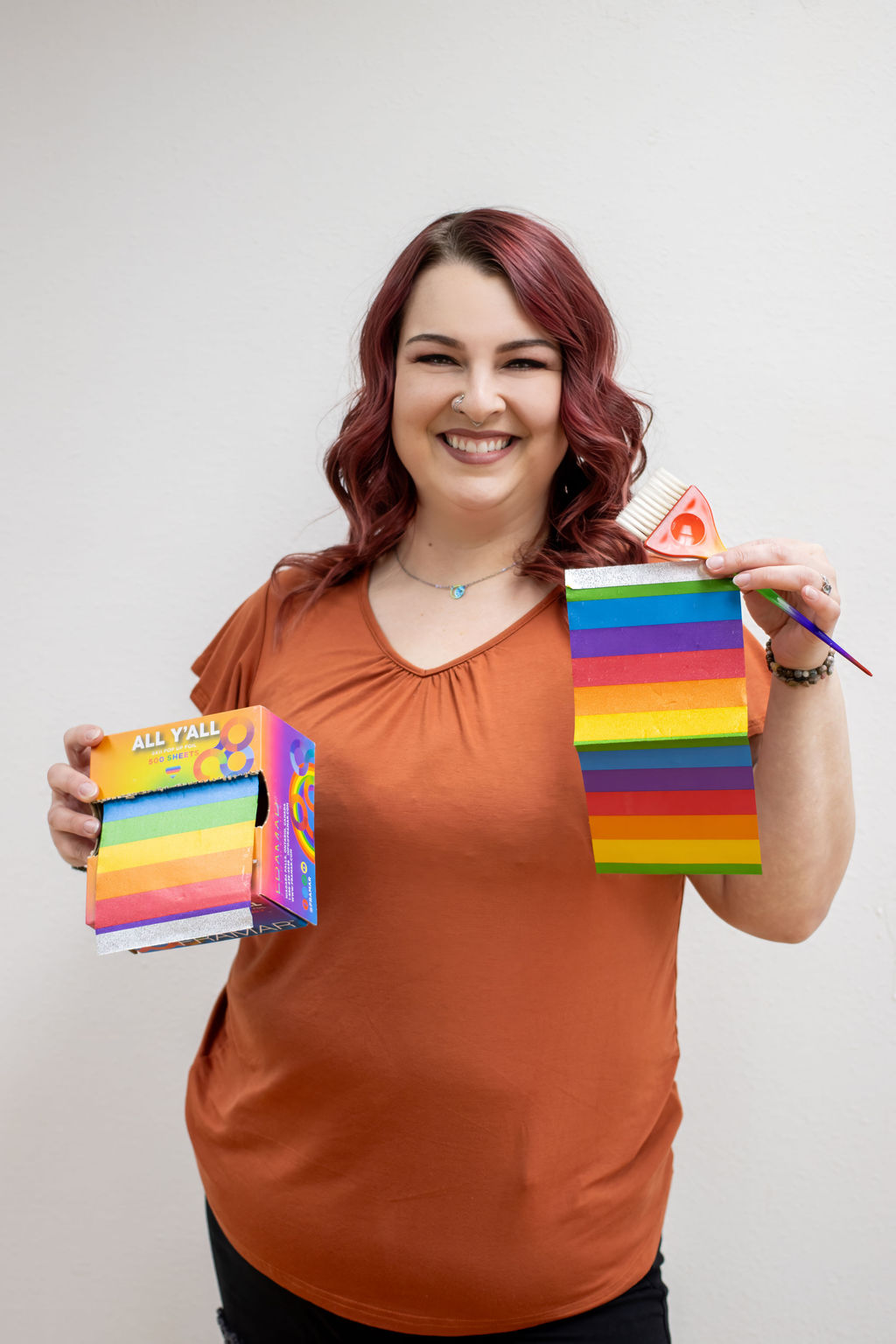 Woman with red hair smiling, holding rainbow-colored sticky notes and a box of sticky notes, wearing an orange top.