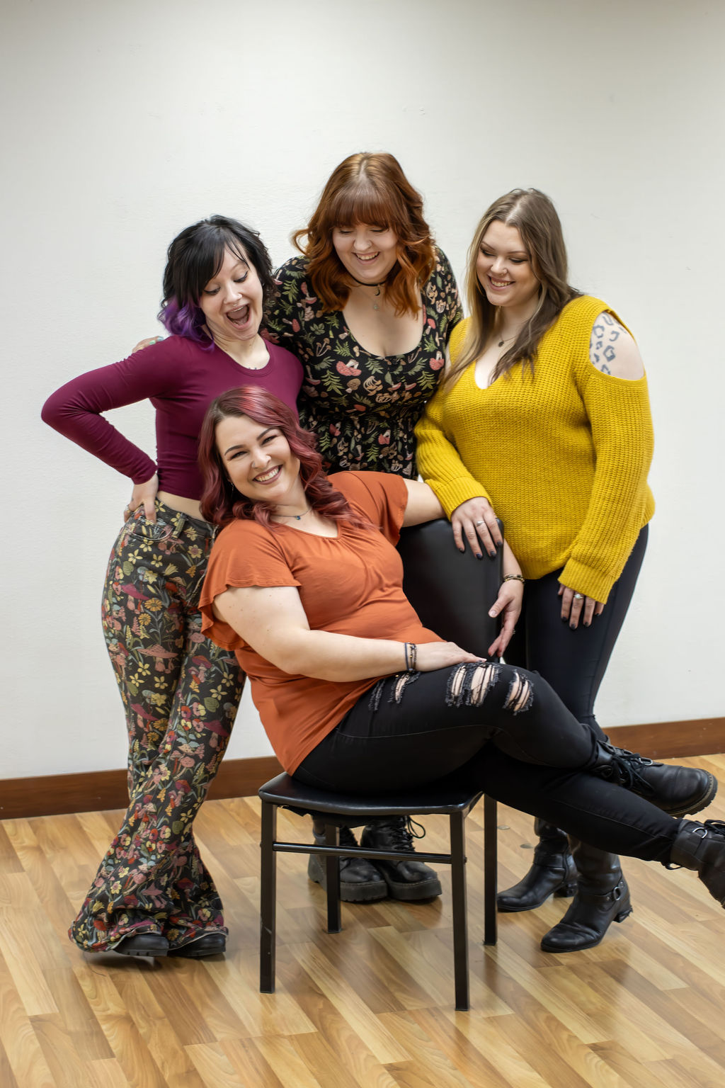Four women smiling and posing together indoors, one sitting on a chair, others standing around her, against a plain wall.
