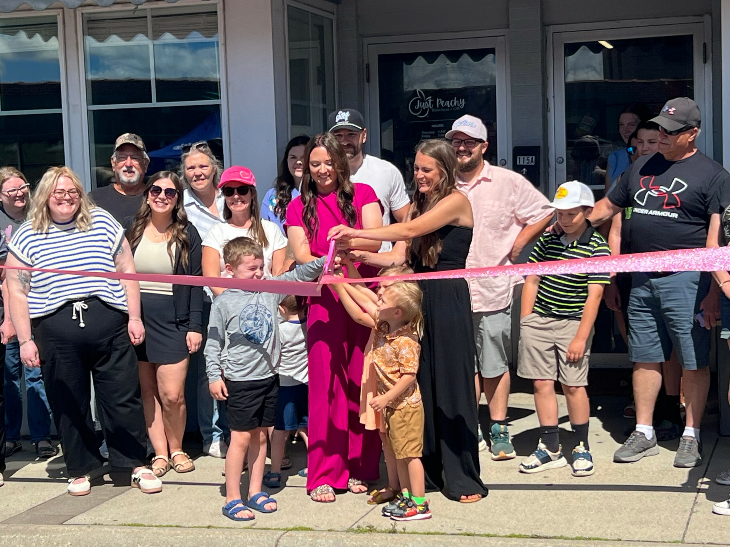 Group of people gathered outside a building for a ribbon-cutting event, with a woman in pink holding scissors, children, and adults smiling.