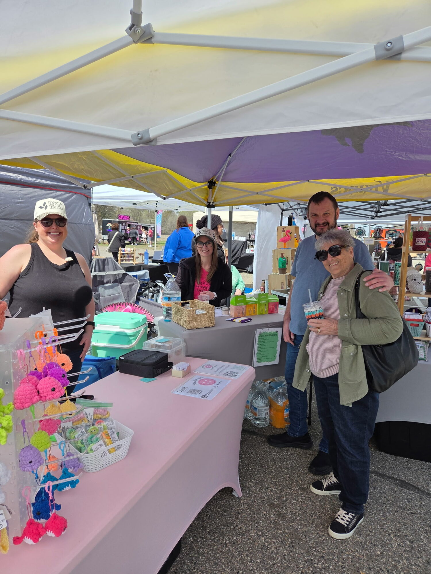 Two women and a man stand under a yellow and purple canopy at an outdoor market stall with colorful items on a pink tablecloth.