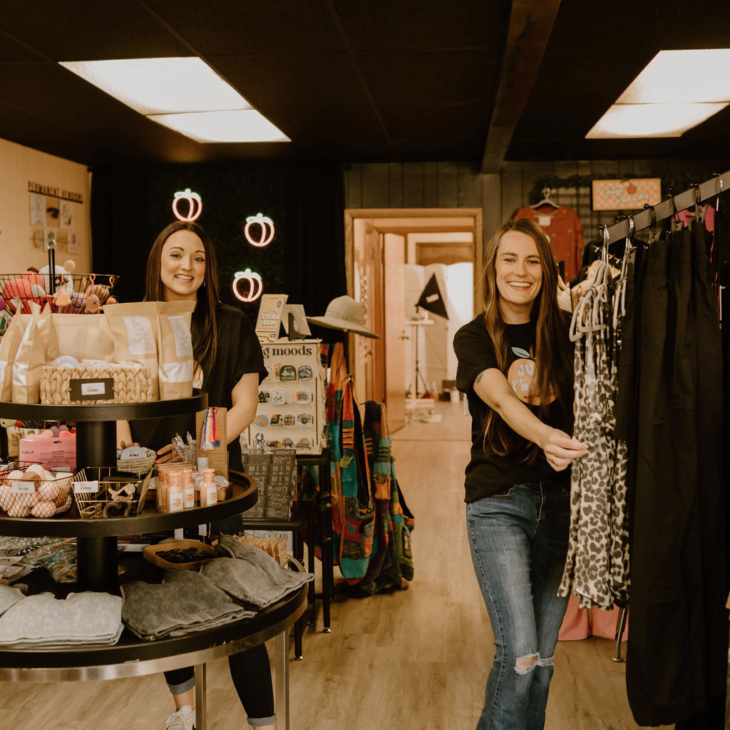 Two smiling women in a boutique store, one behind a display and the other browsing clothes, with warm lighting and clothing racks.