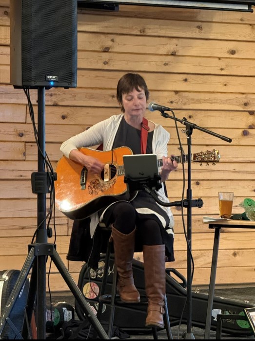 Person playing guitar and singing into a microphone, seated indoors with wooden wall background, a table with a drink nearby.