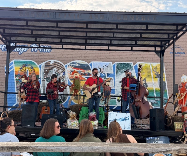 Group of musicians performing on an outdoor stage with a colorful mural background, audience watching, and a sign reading 'SLO COUNTY FAIR'.