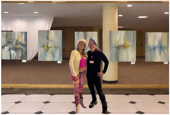Two women stand in front of abstract paintings in an art gallery, smiling and posing for the photo.
