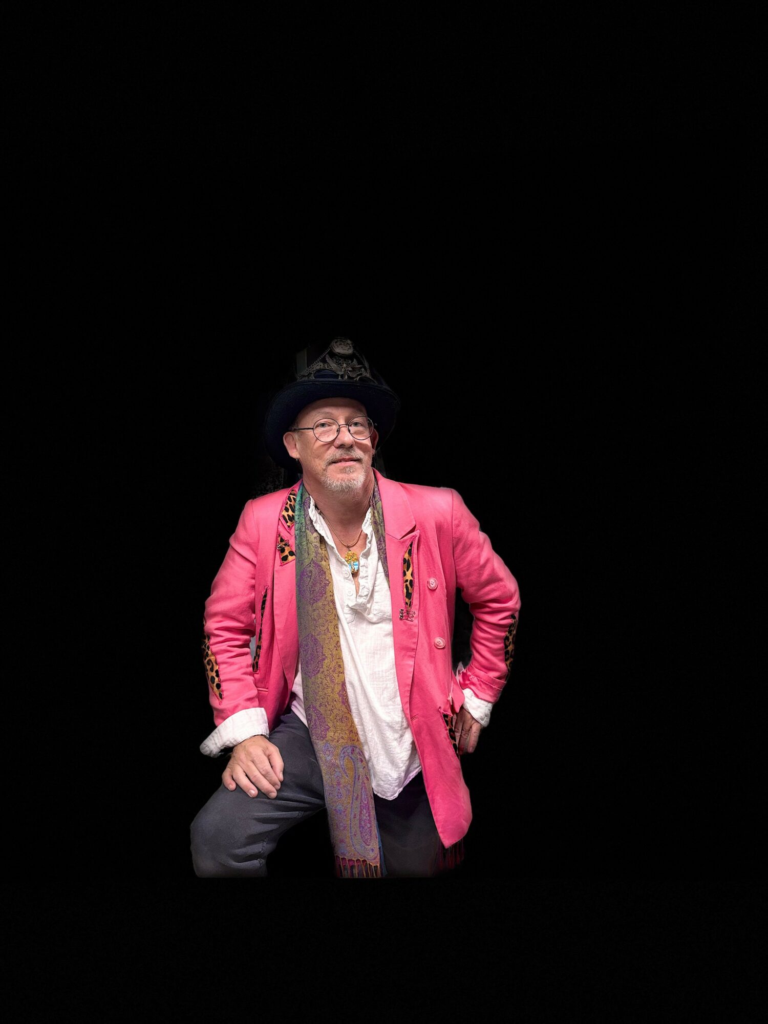 Person wearing a pink jacket, black hat, glasses, and jewelry, standing against a black background.