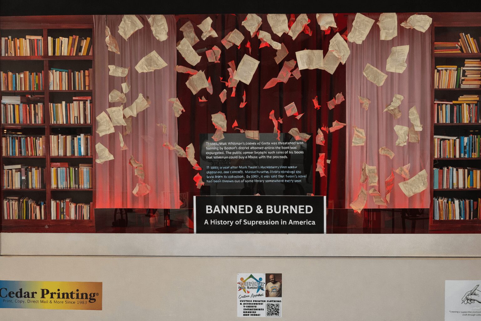 Bookshelves with books, a stage with curtains, and papers falling, with a sign reading 'BANNED & BURNED' and a subtitle.