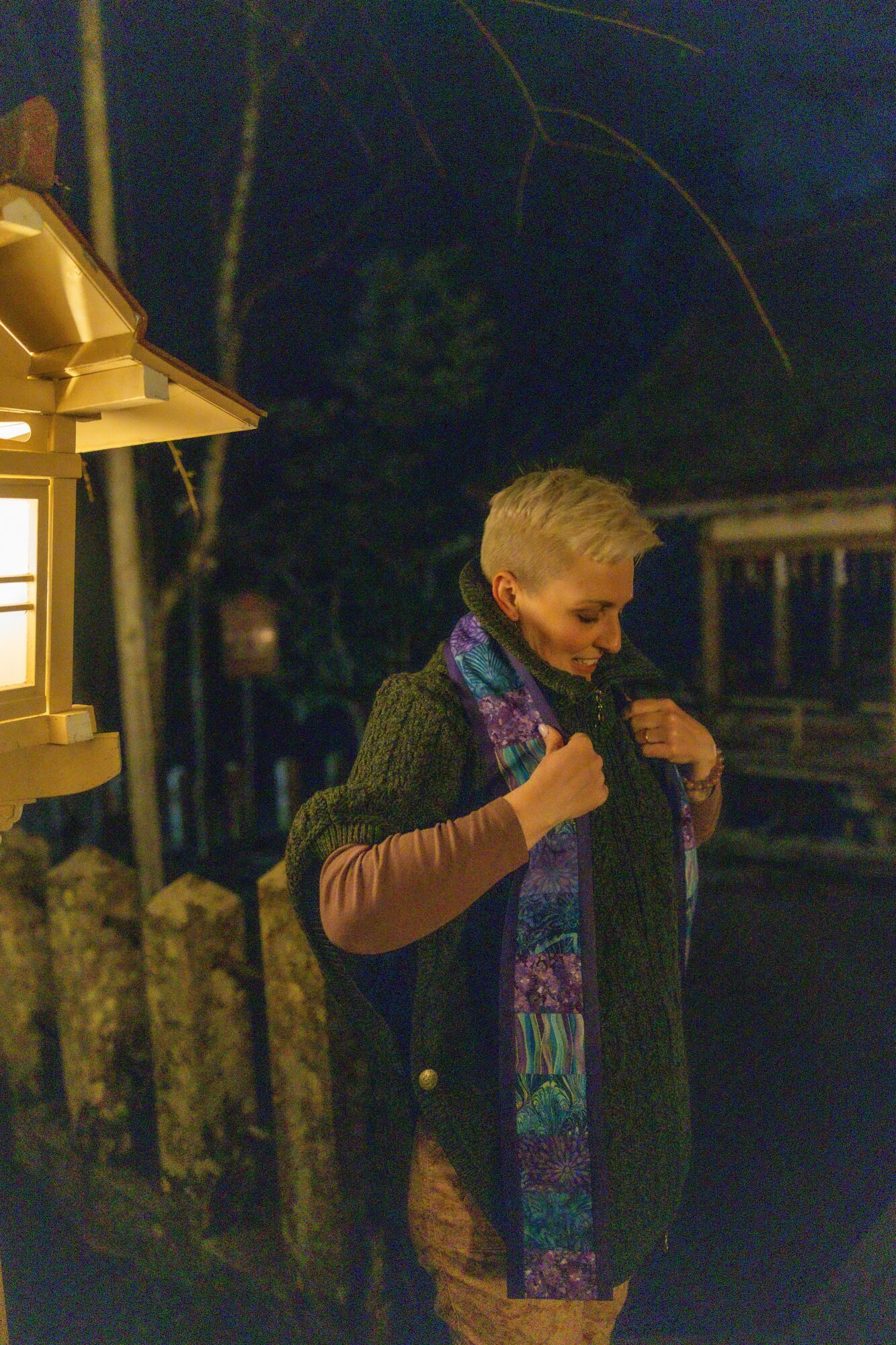 Woman standing outdoors at night, wearing a scarf and coat, near a lit lamp post and wooden fence.