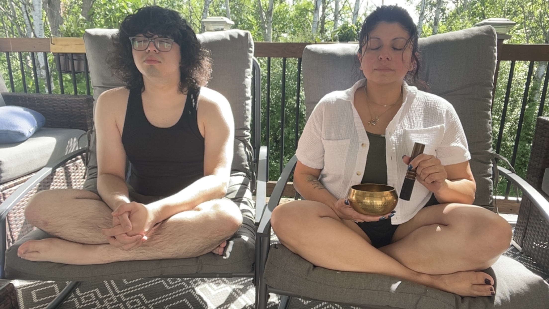 Two women sit cross-legged on outdoor chairs, holding bowls, with trees and a wooden railing in the background.