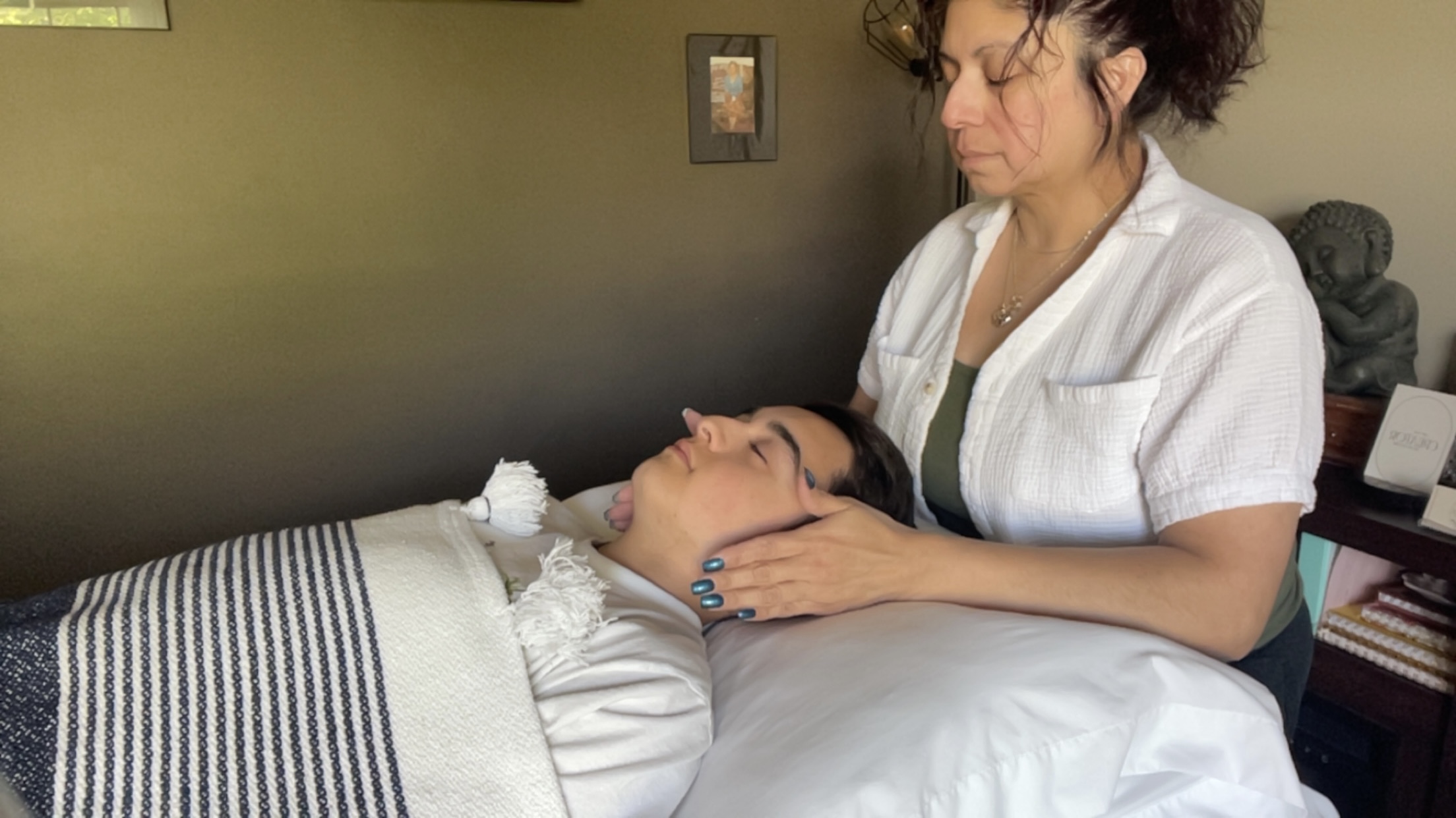 A woman in a white uniform gently holds a person's face during a massage or therapy session in a room with a dark wall and framed picture.