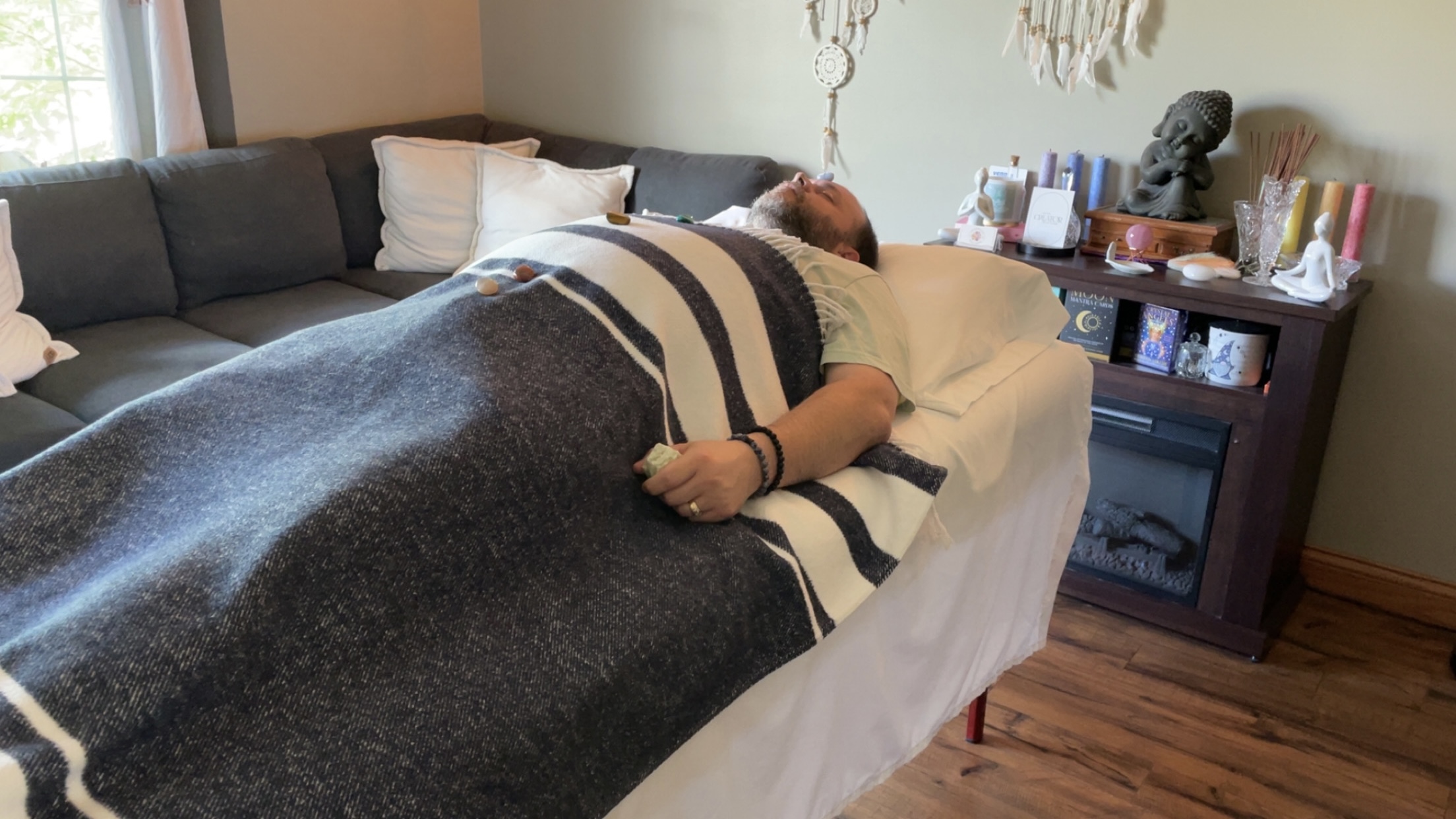 Person lying on a sofa covered with a blanket, in a living room with a side table and books.