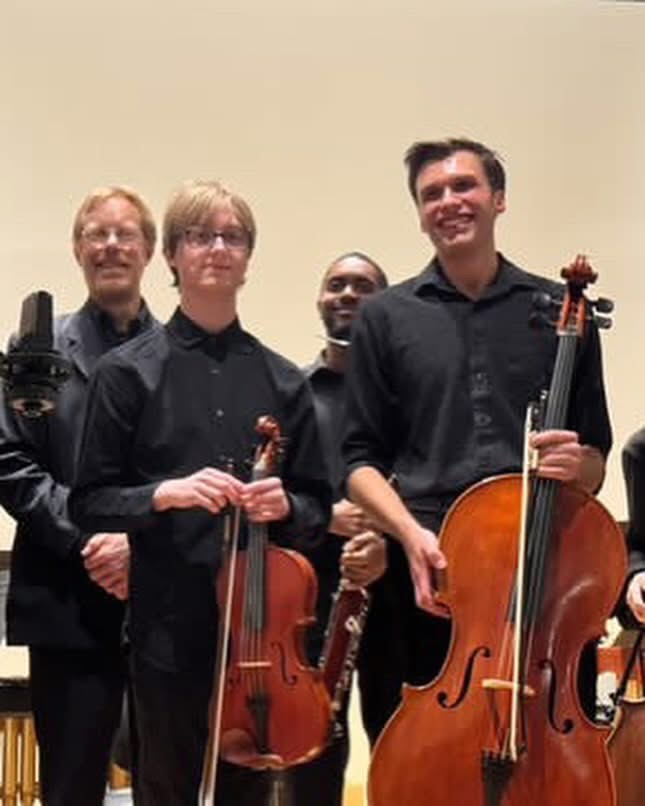Four musicians holding string instruments, smiling, standing against a plain background.
