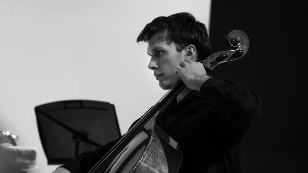 Young man playing a cello, focused, in a room with a music stand, black and white photo.