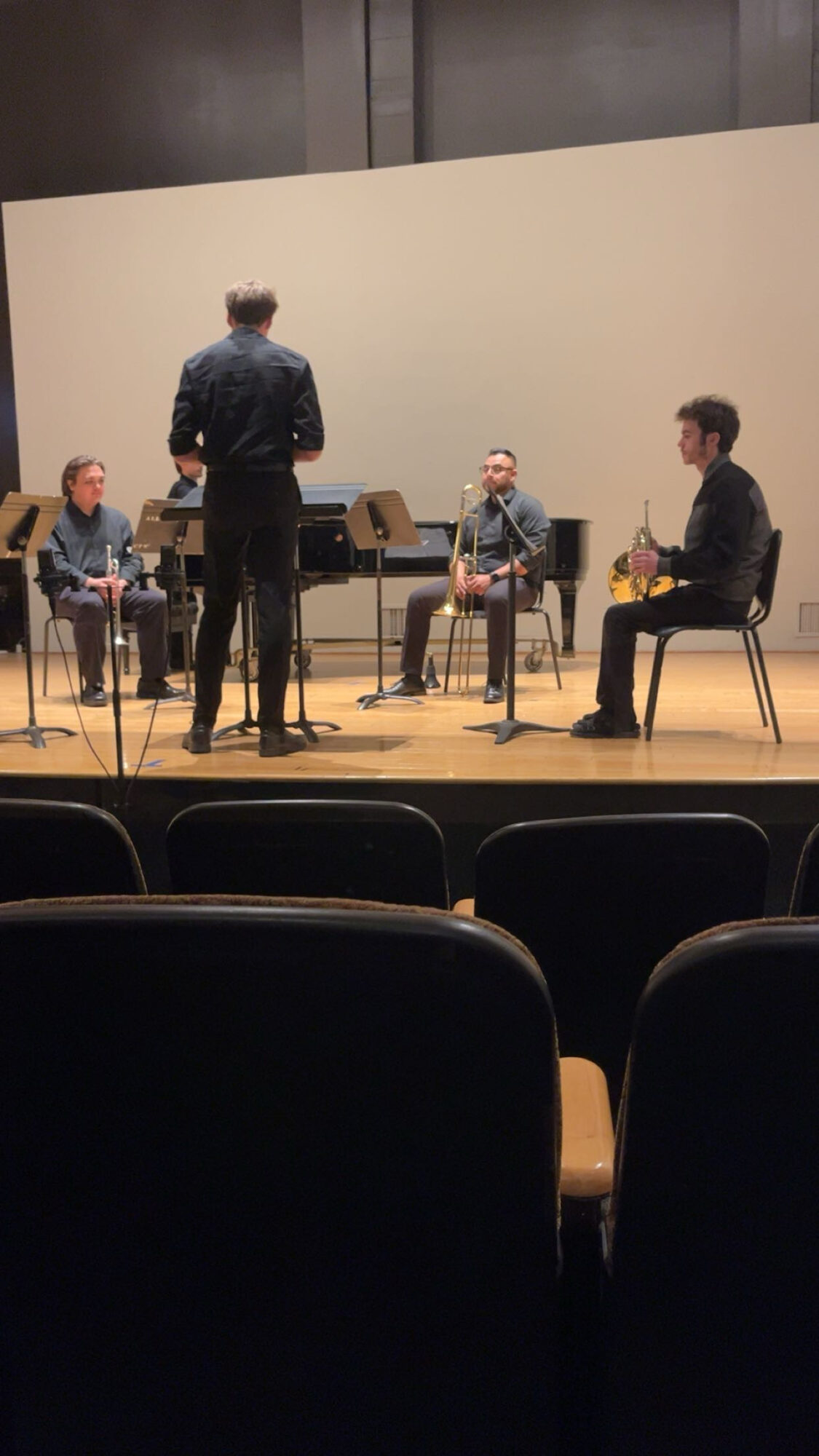 Four people on stage, three seated and one standing, with chairs and a plain background, in a theater setting.