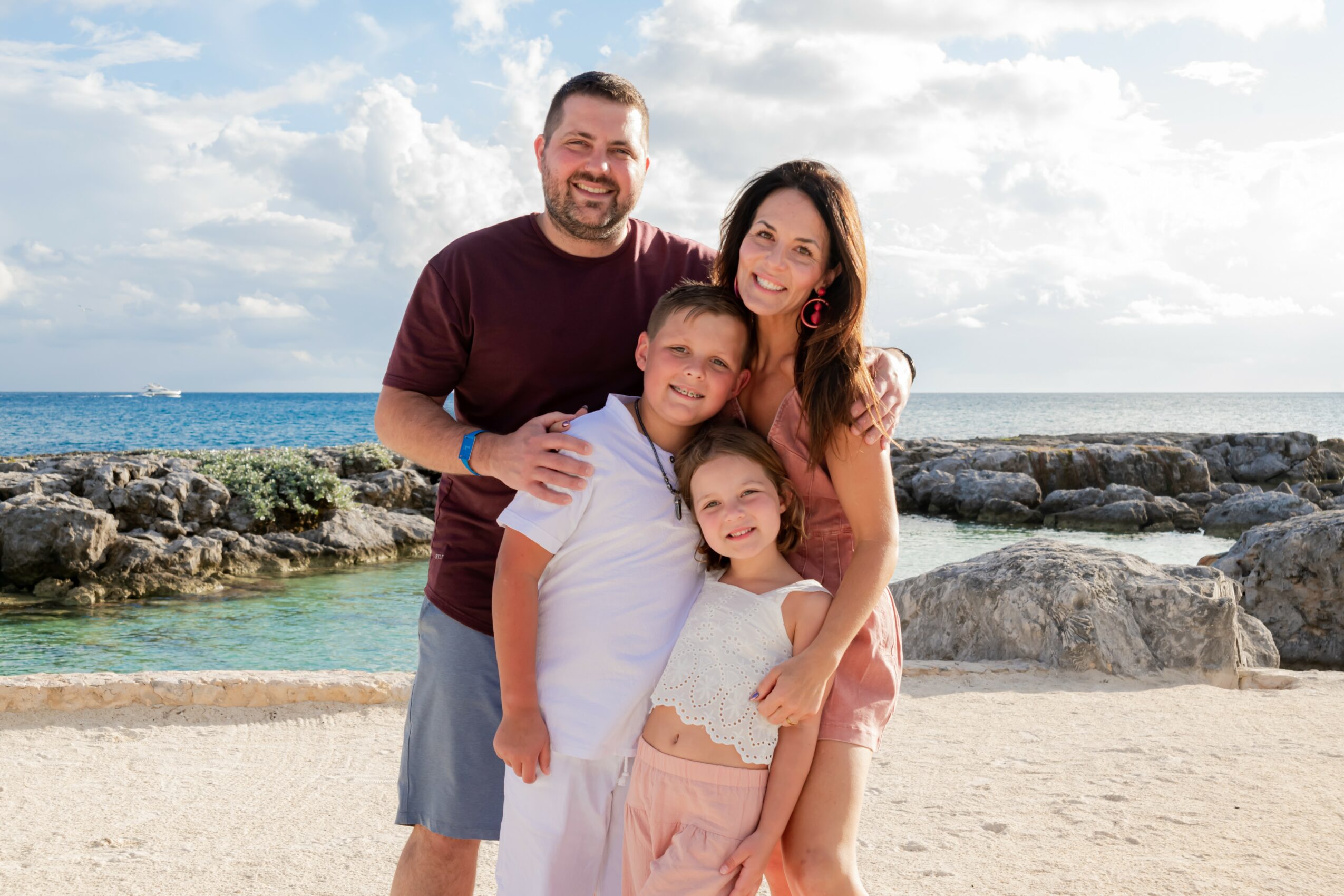 Family of four standing on a beach with rocks and ocean in background, smiling, sunny sky with clouds.