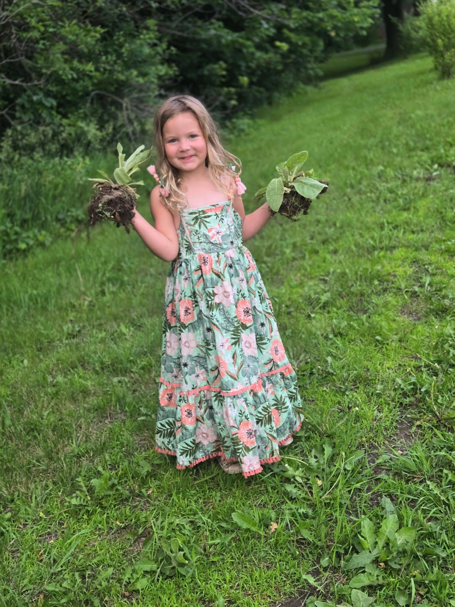 Young girl in a floral dress holding plants in a grassy outdoor area.