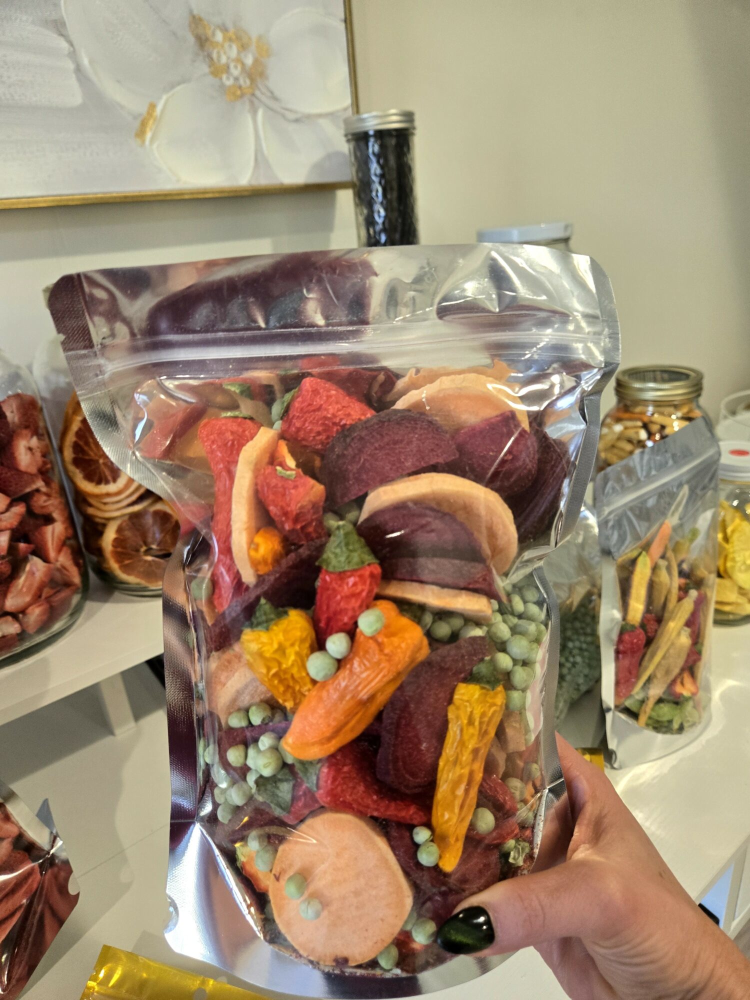 Hand holding a clear plastic bag filled with colorful candies and chocolates, on a white surface with jars and a flower painting in background.