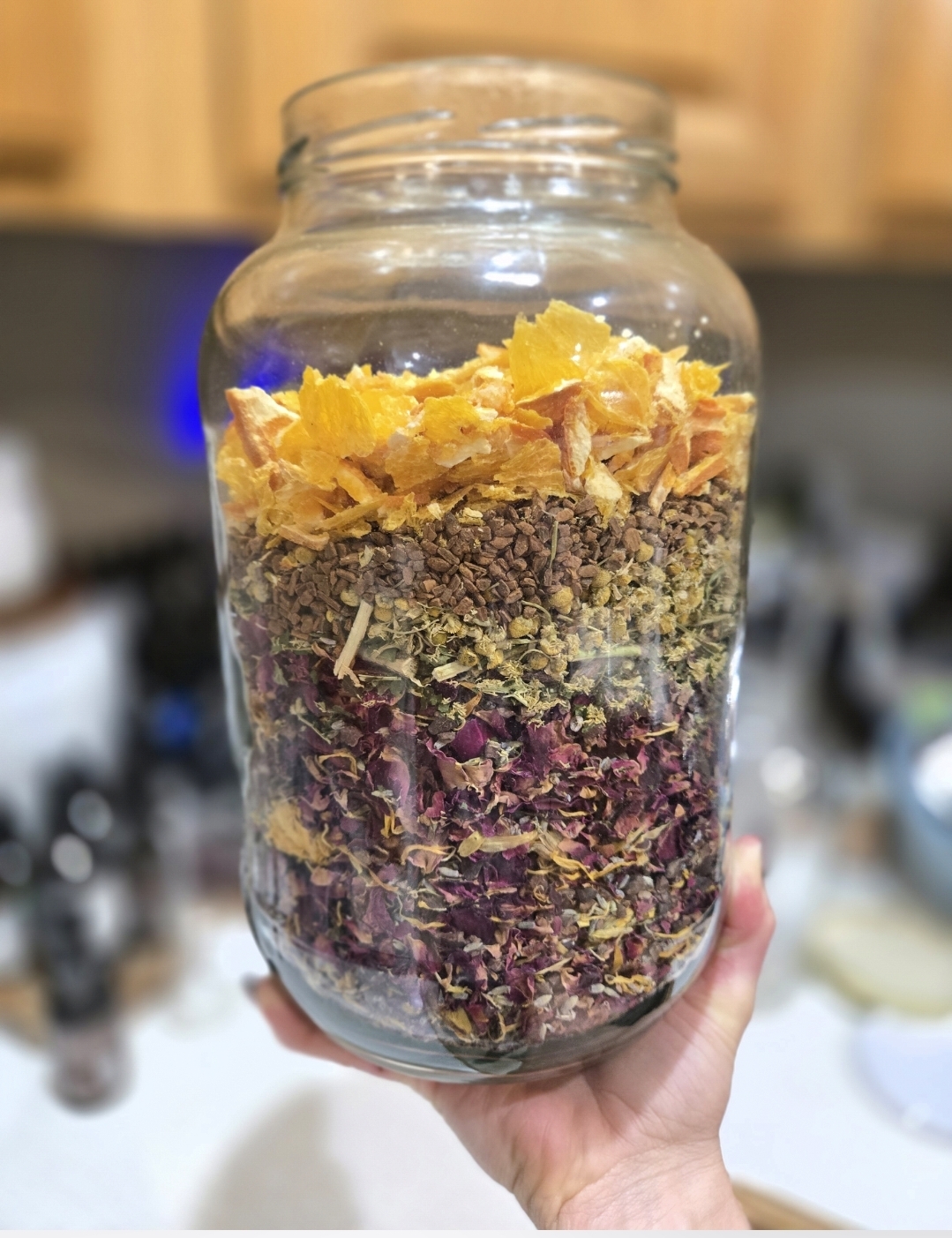 Hand holding a glass jar filled with layered dried herbs and flowers, kitchen background, clear focus.