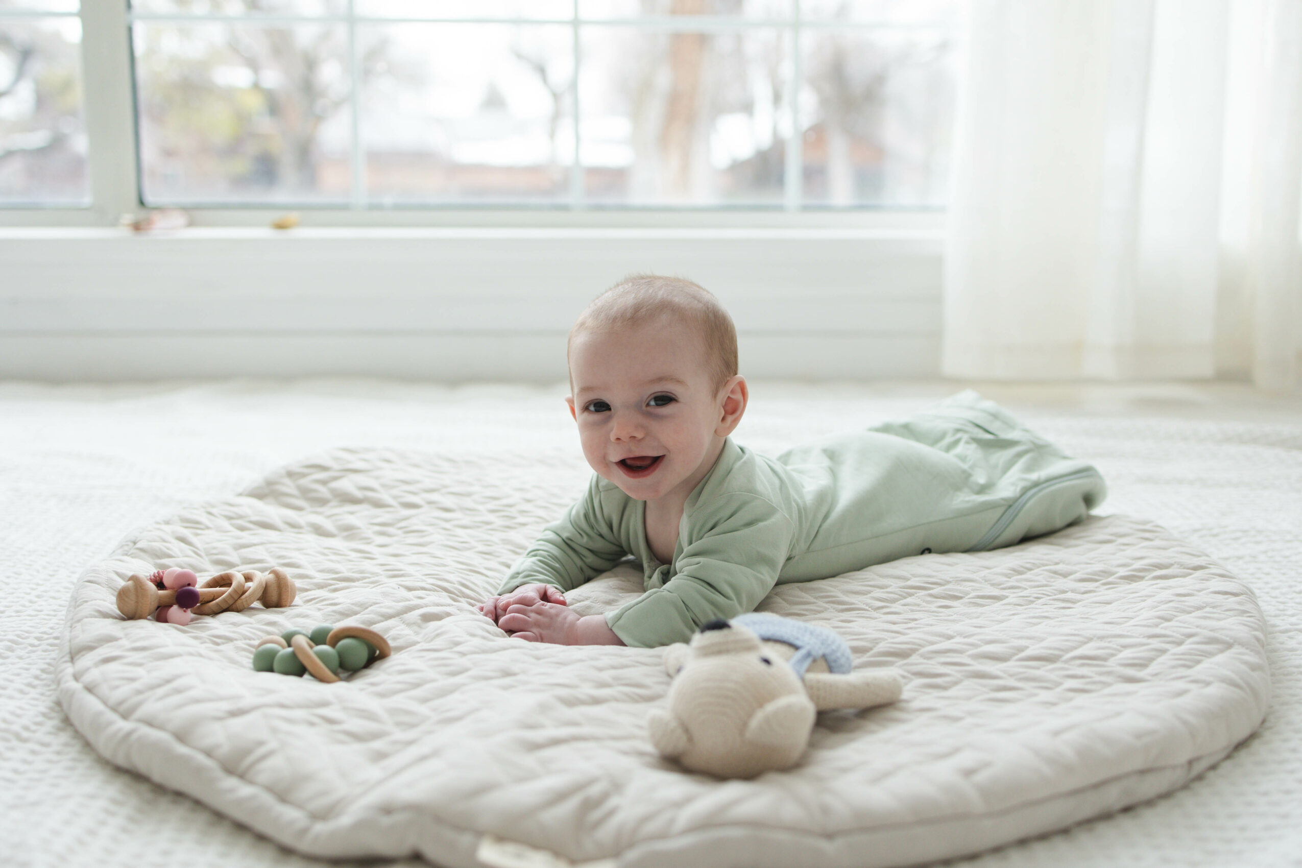 Baby lying on a quilted mat with toys, smiling, in a bright room with large windows and curtains.