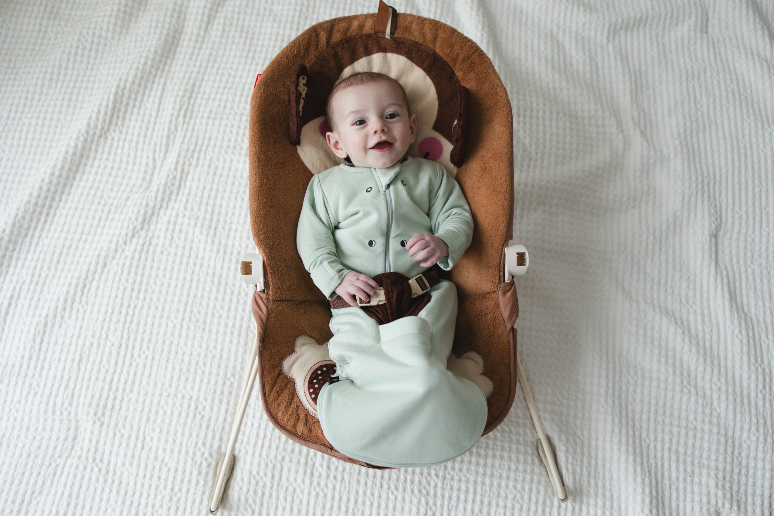 Baby lying in a brown cushioned baby rocker on a white surface, smiling and looking up.