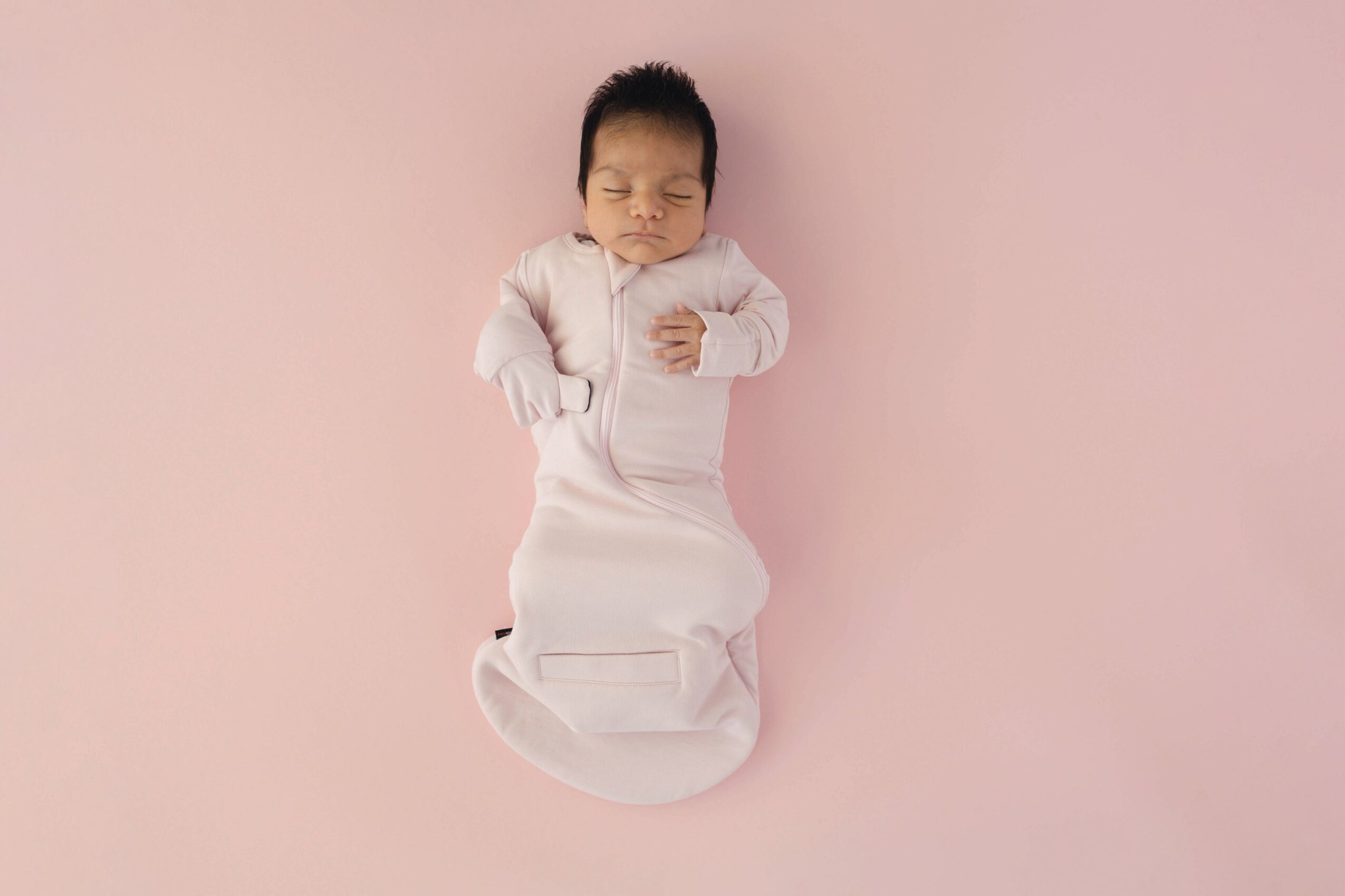 Baby lying on pink background, wearing a white outfit, with eyes closed and hands near face.