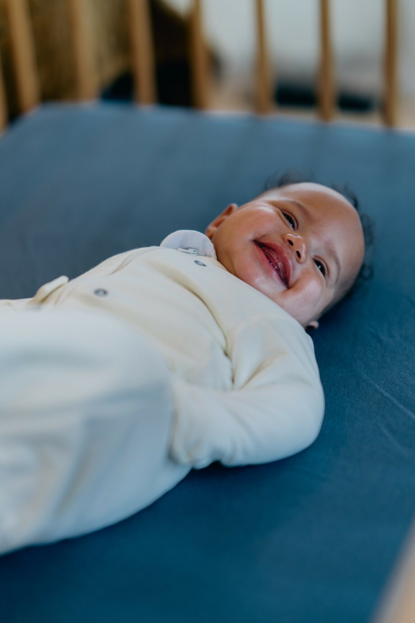 Baby lying on a bed, smiling and looking up, wearing a white outfit, with a wooden headboard in the background.