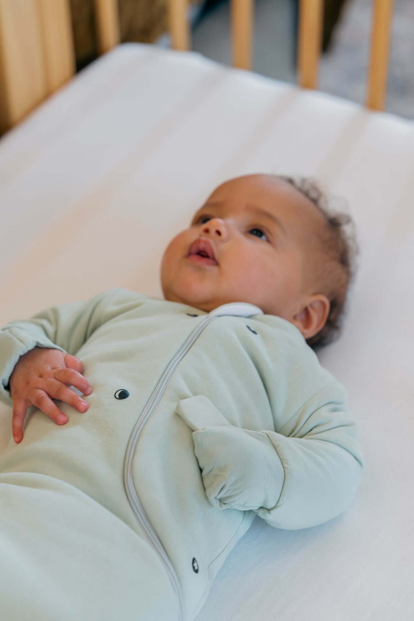 Baby lying on a bed, looking upward, wearing a light-colored onesie, with a wooden headboard visible in the background.