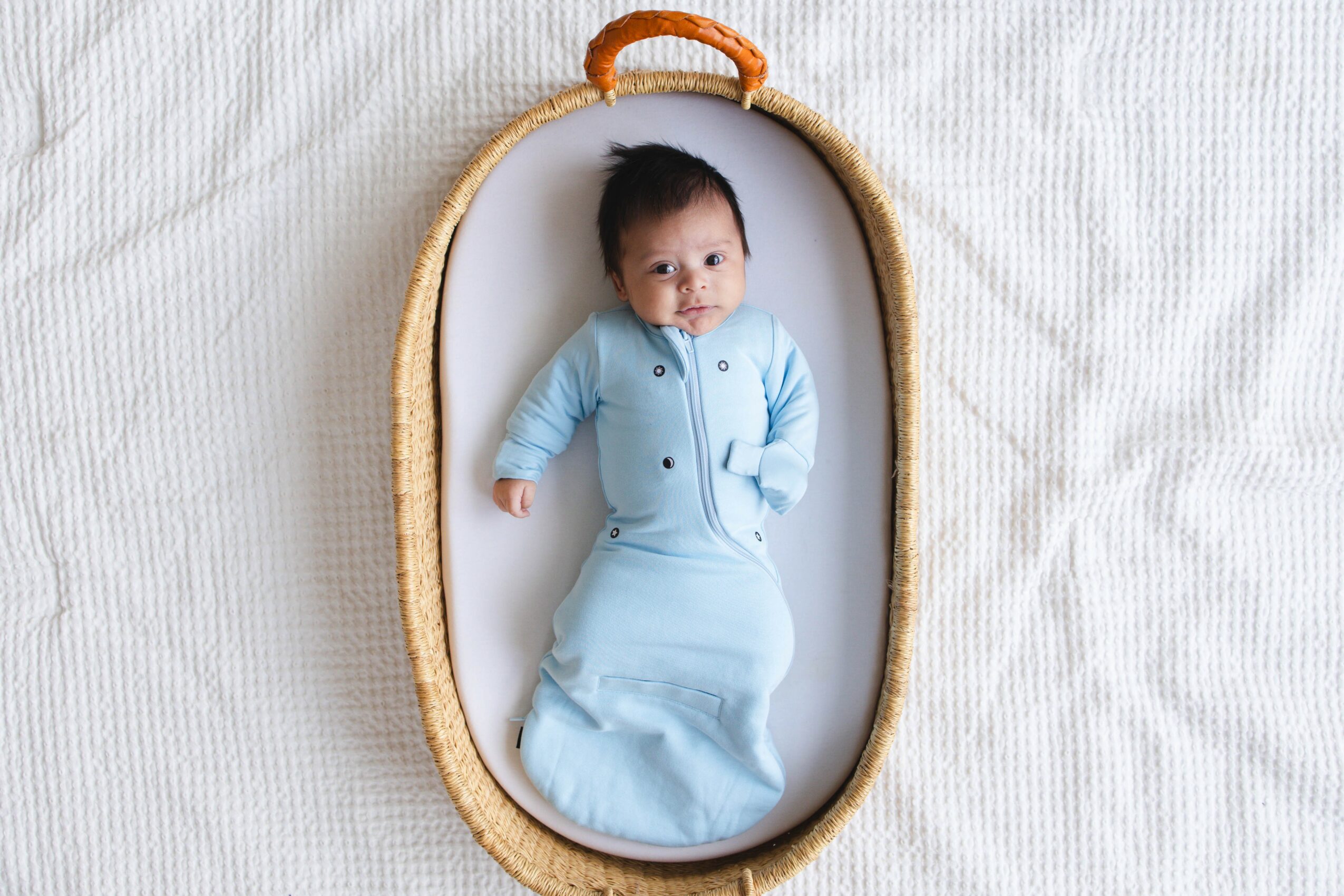 Baby lying in a woven basket with a white background, wearing a light blue outfit, looking at the camera.