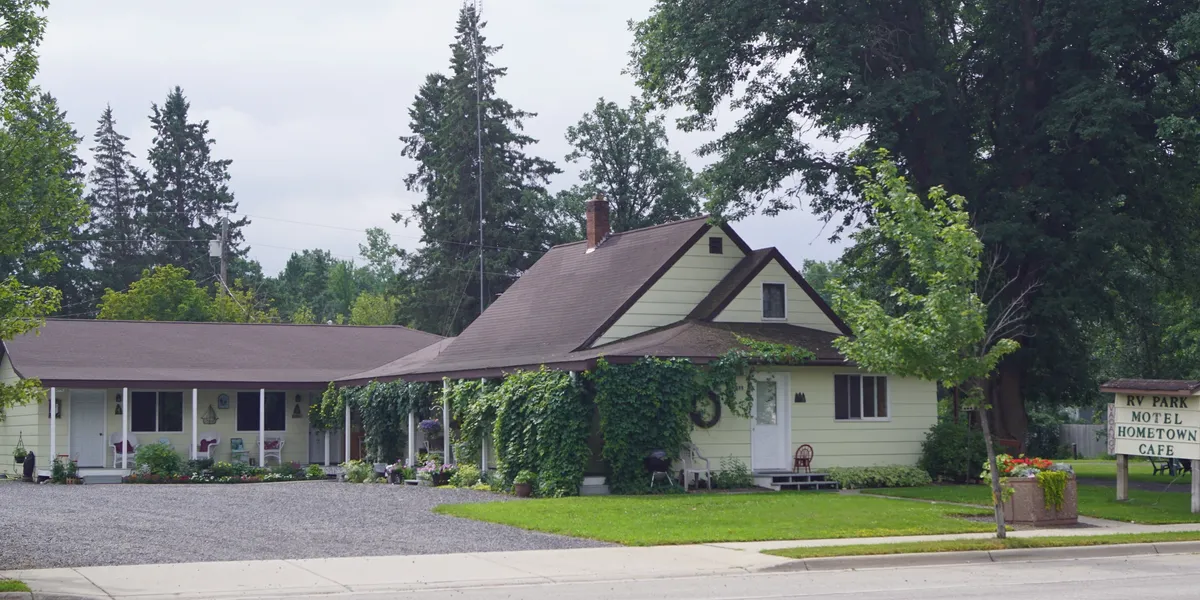 A house with a sloped roof, surrounded by trees and a lawn, with a gravel driveway and a signpost.
