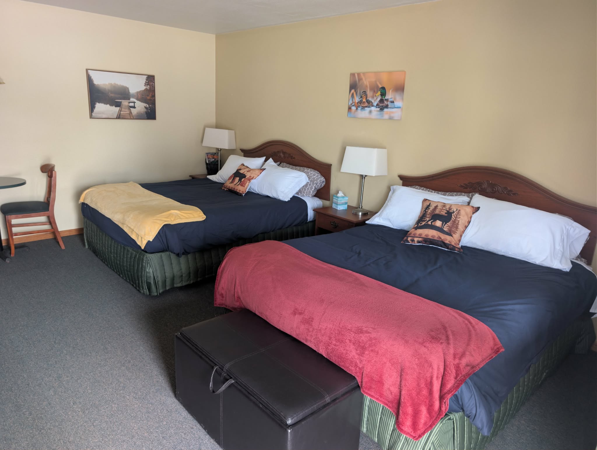Two beds with pillows and blankets, bedside lamps, a chair, and framed pictures on the wall in a hotel room.