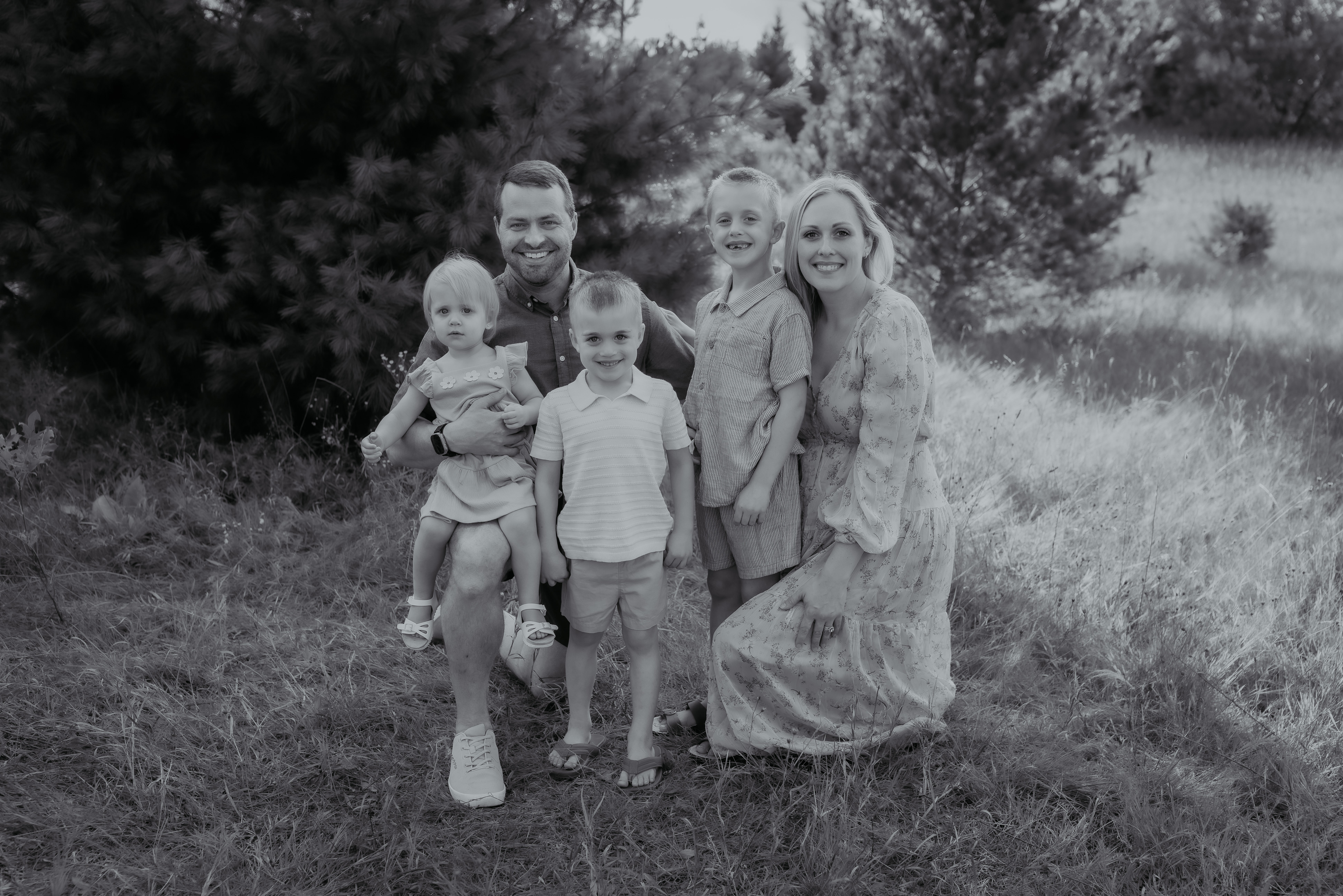 Group of six people outdoors, standing on grass with trees in background, smiling at camera.