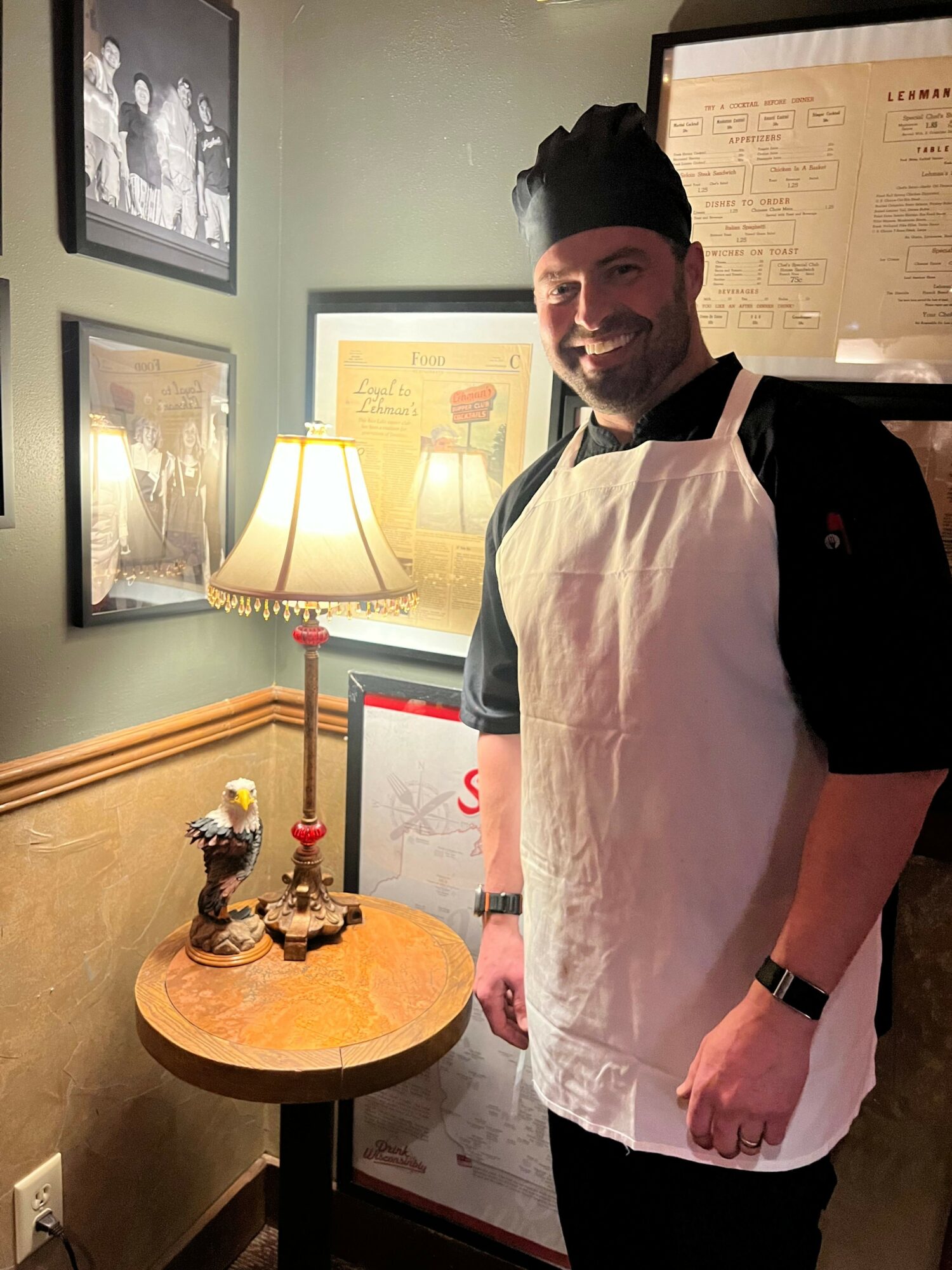Person standing indoors near framed pictures, wearing a white apron, black shirt, and a watch, with a table and lamp nearby.