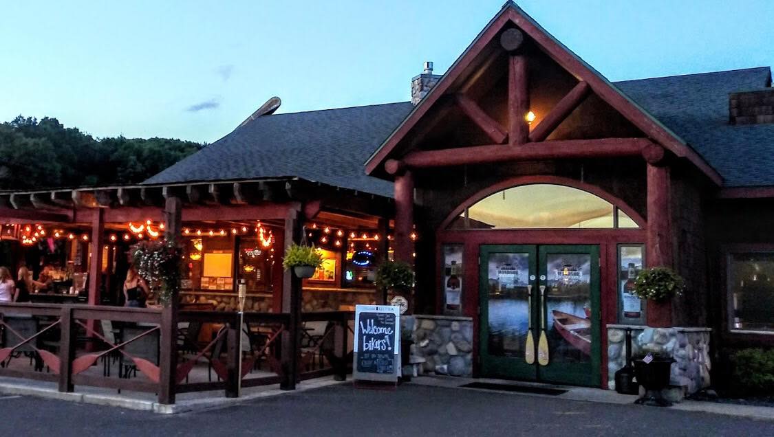 Exterior of a rustic building with a bar area, outdoor seating, and a sign, illuminated at dusk.