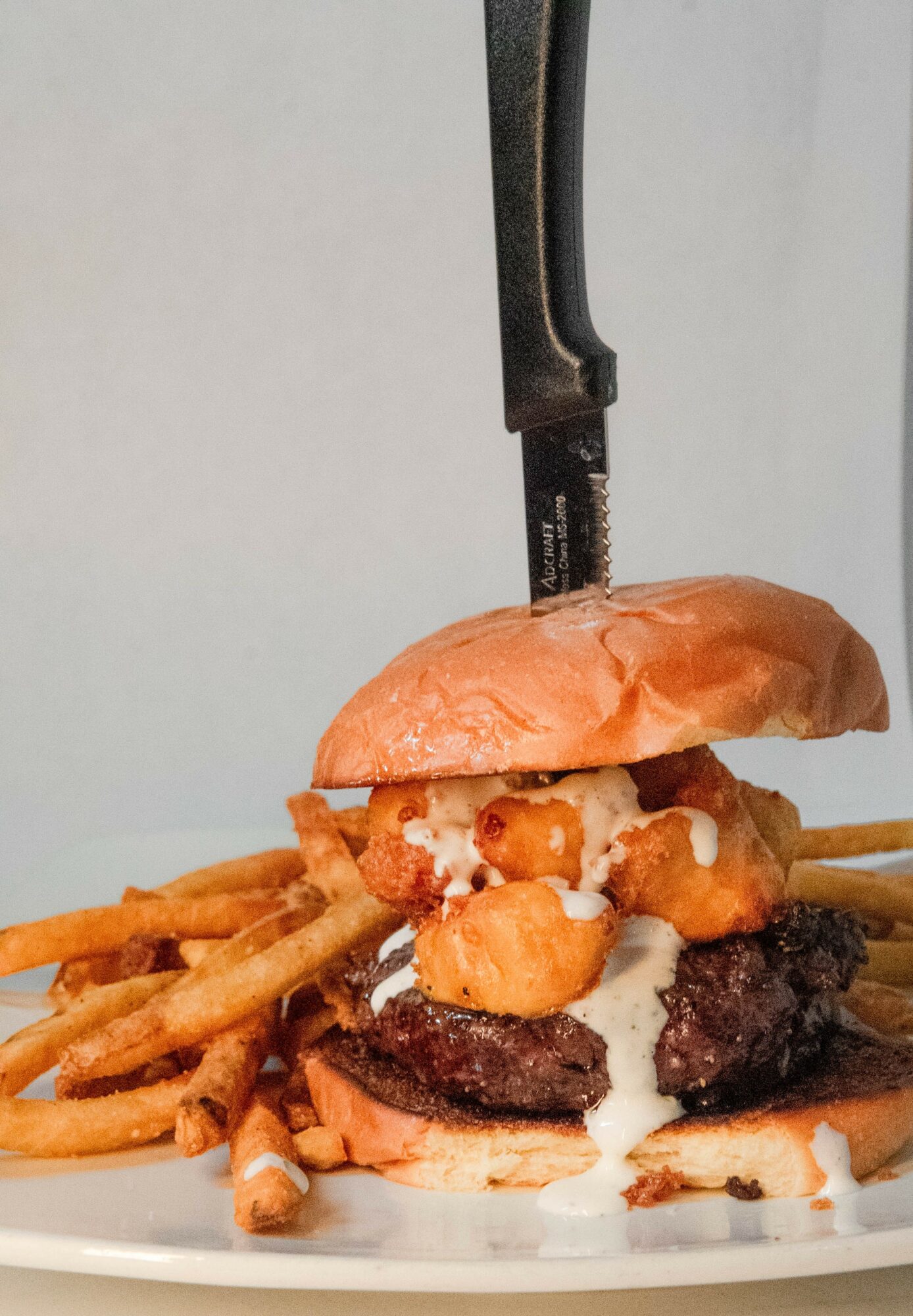 A burger with fries and sauce on a plate, with a knife inserted through the top bun, against a plain background.