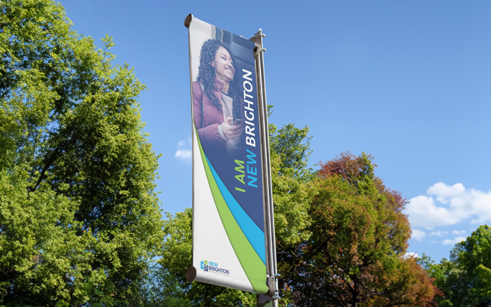 Flag with a woman and text 'I AM NEW BRIGHTON' against a blue sky with trees.